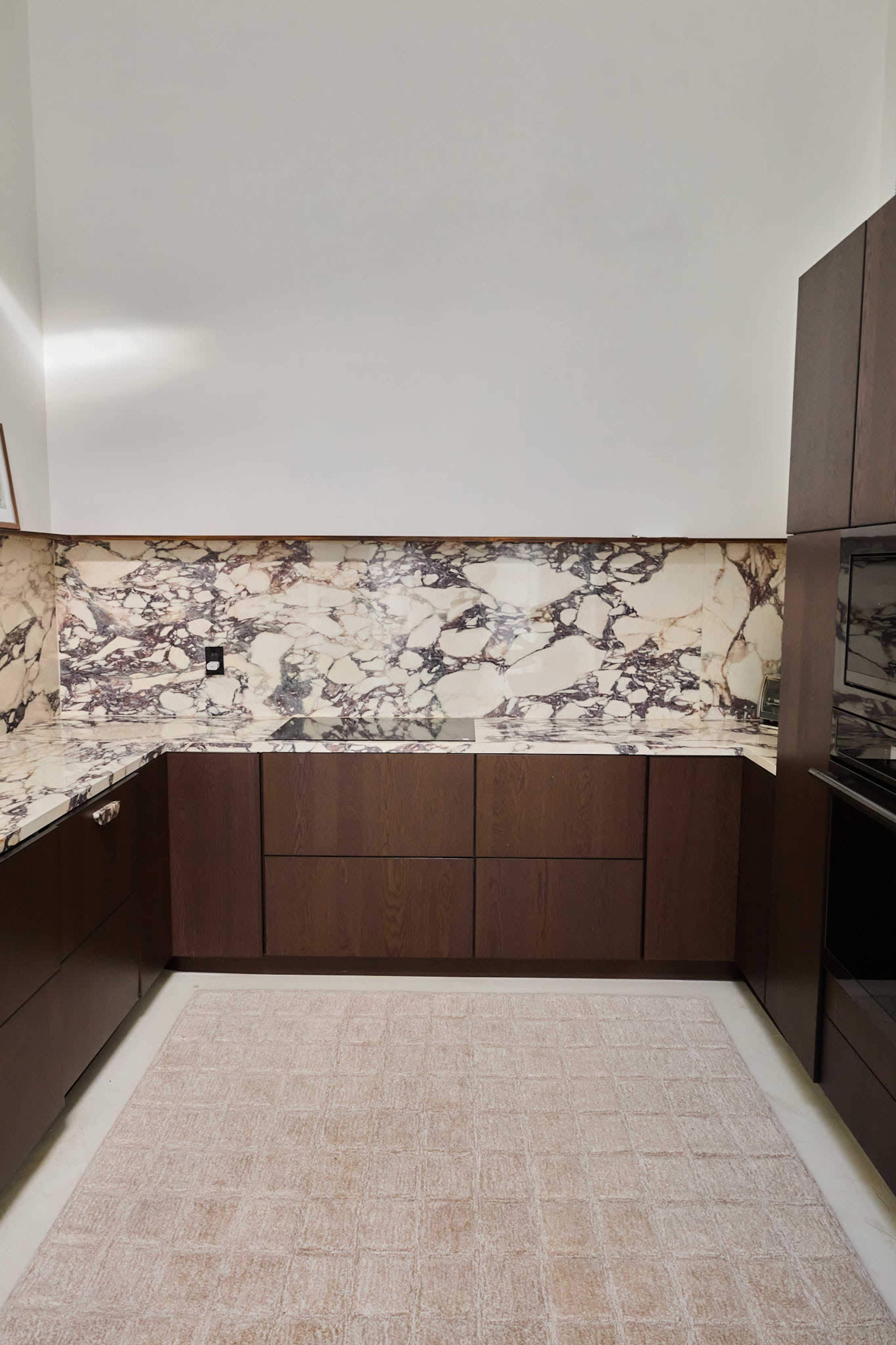 The image shows a modern kitchen with dark wooden cabinetry and a marble backsplash featuring a mix of cream and brown tones, alongside a light-colored rug on the floor.