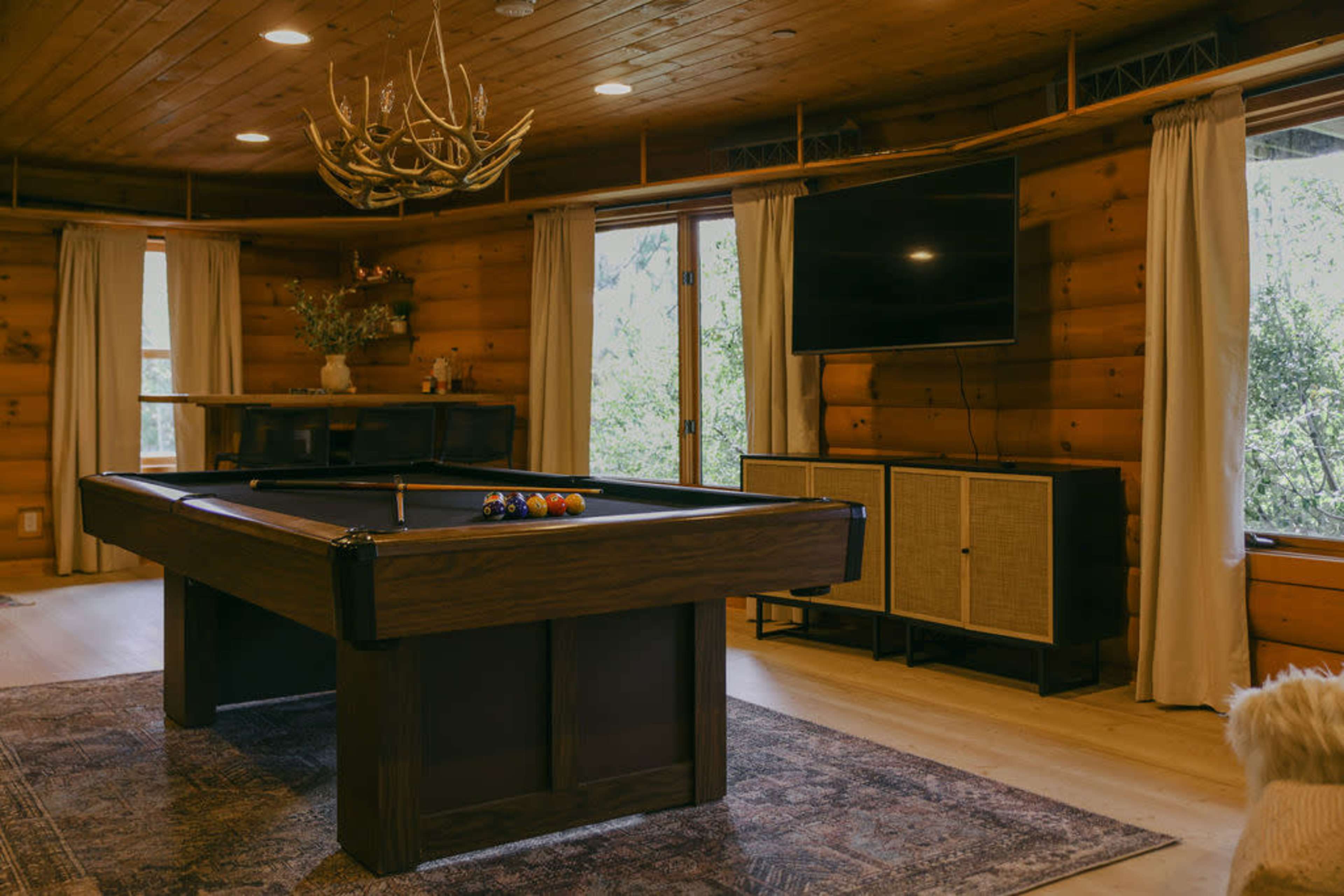 The image shows a rustic game room featuring a wooden pool table, a flat-screen TV mounted on the wall, and large windows with natural light illuminating the space.