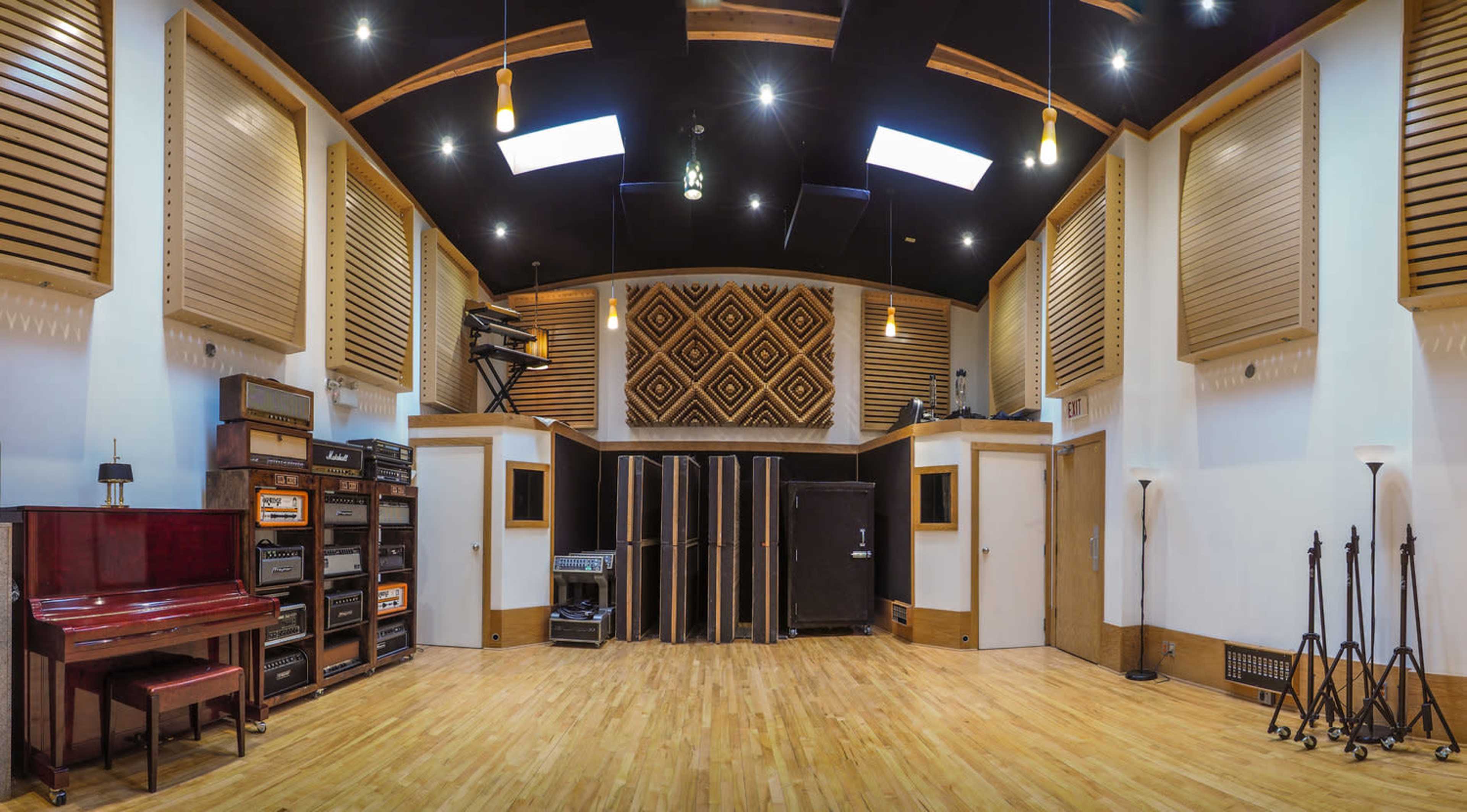 A spacious music studio with a wooden floor, soundproof walls, various amplifiers, a piano, and overhead lighting.