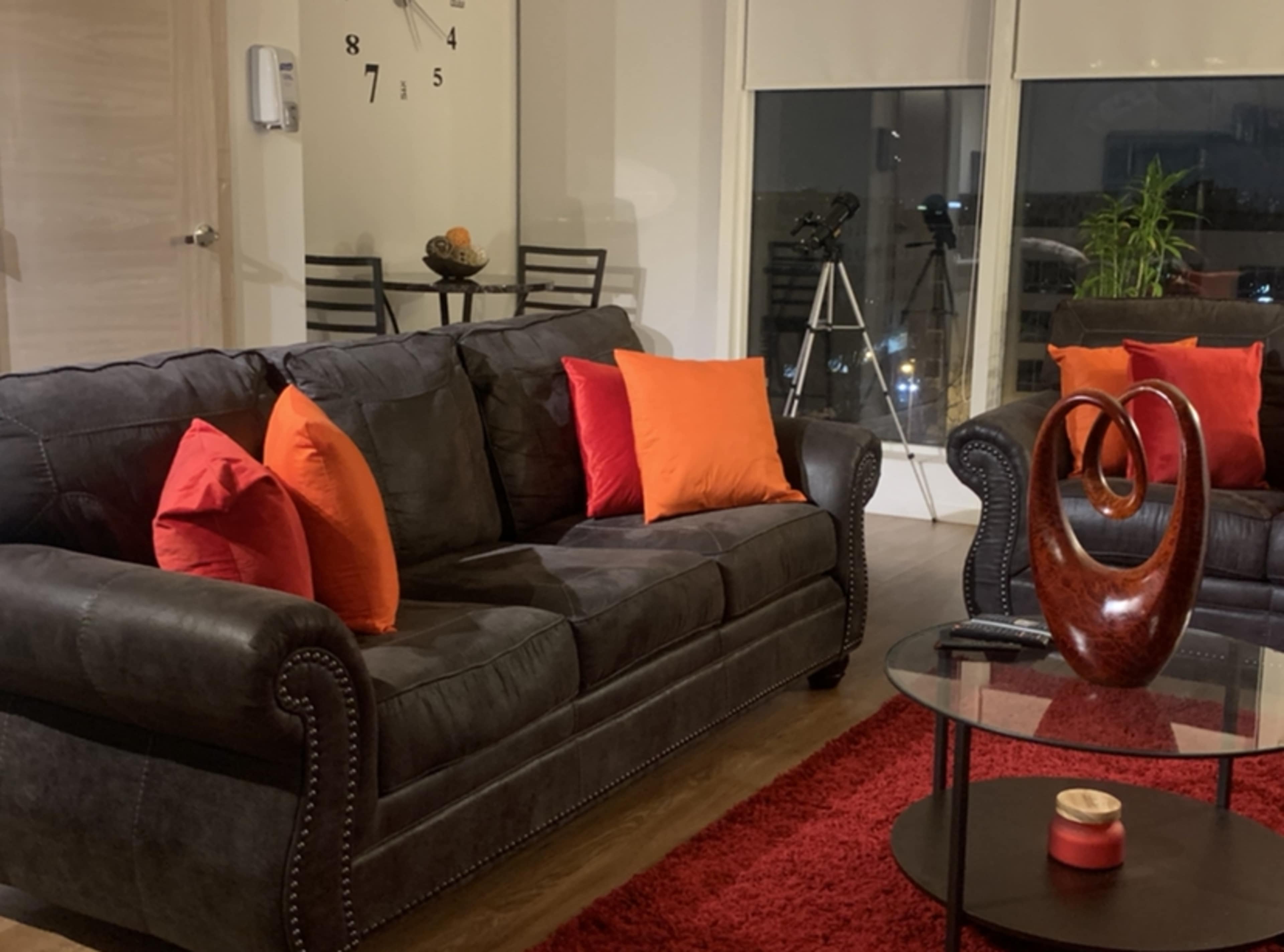 A black sofa adorned with orange pillows sits in a modern living room, complemented by a round glass coffee table and a tripod telescope in the background.