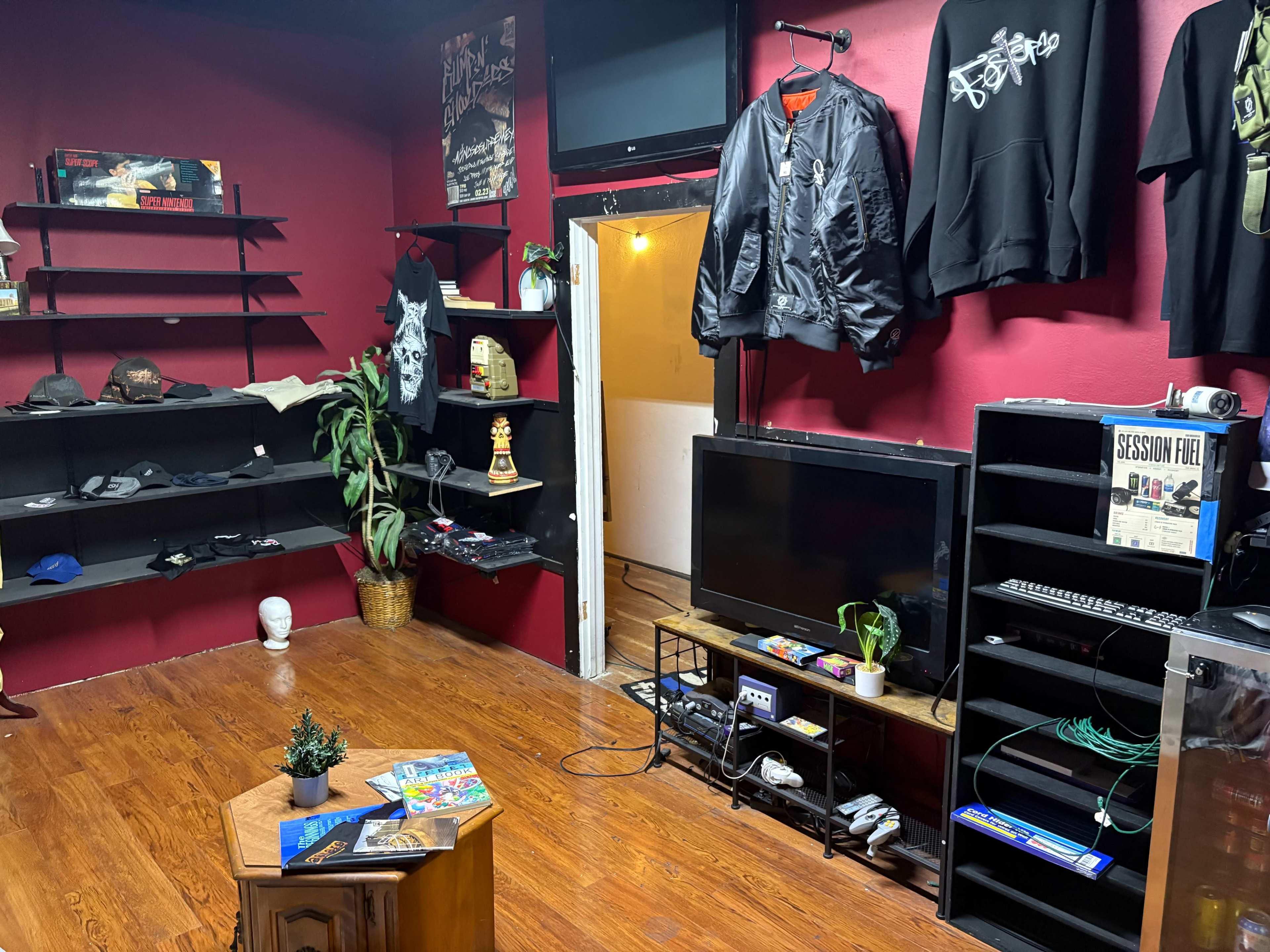 The image shows a room with red walls, a television and a shelf filled with clothing and various items, alongside a small table in the center and a doorway at the back.