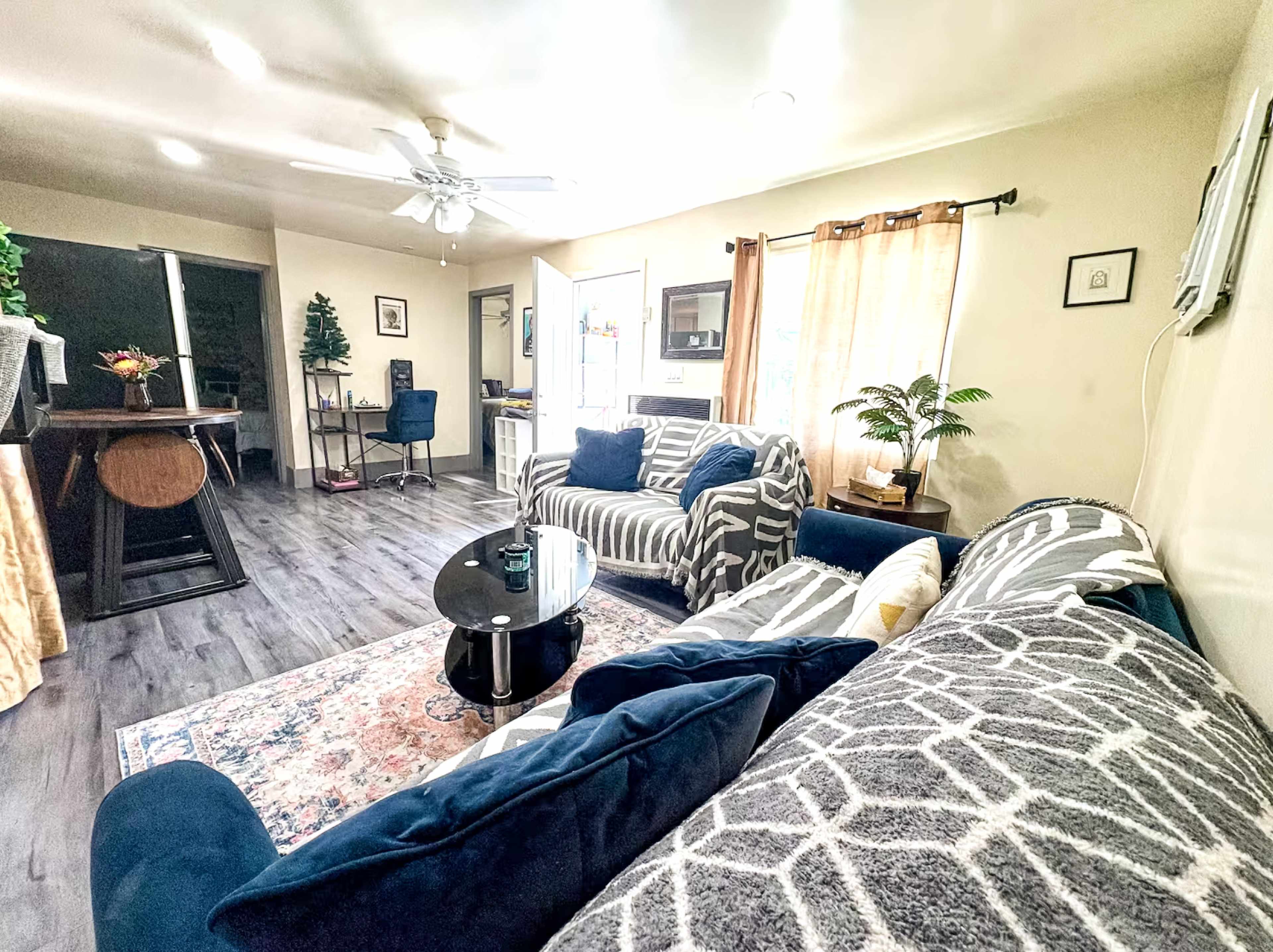 A bright living room features two patterned sofas, a black coffee table, and a view of a workspace area with a desk and chairs.