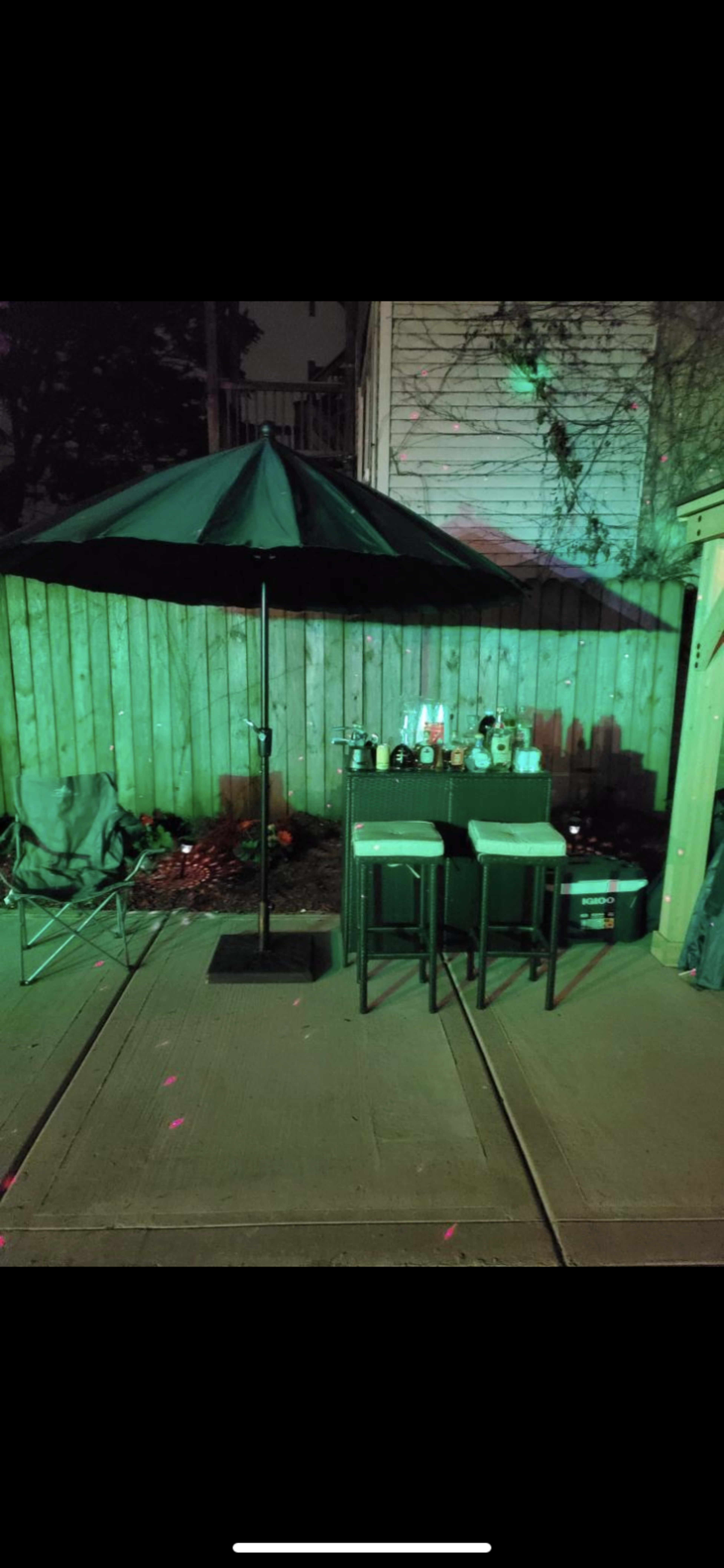 A patio setup features a green umbrella, two stools, and a bar area with various bottles against a wooden fence.