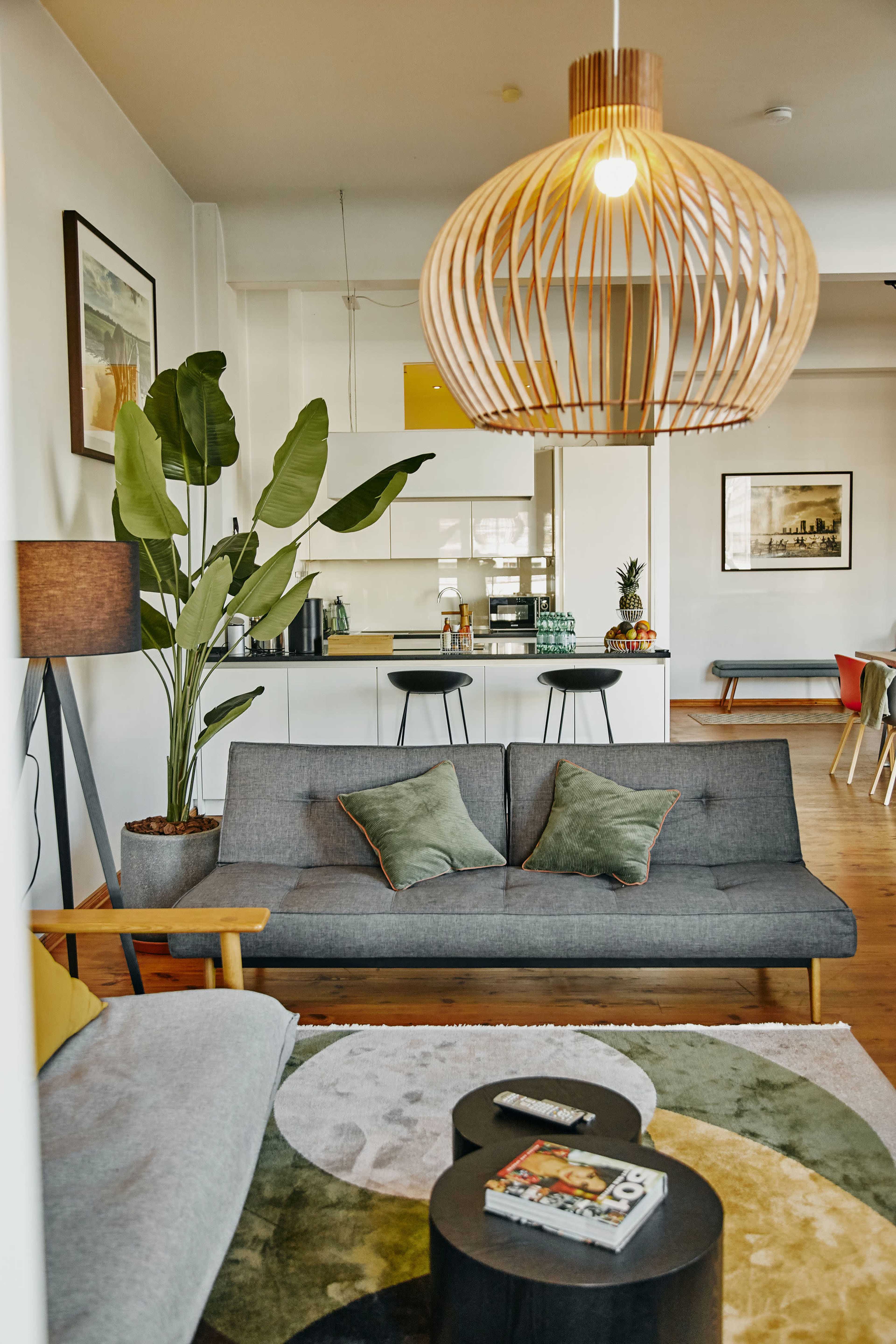 The image shows a modern living room with a grey sofa, a round coffee table, a large plant, and a pendant light above, leading into a bright kitchen area in the background.