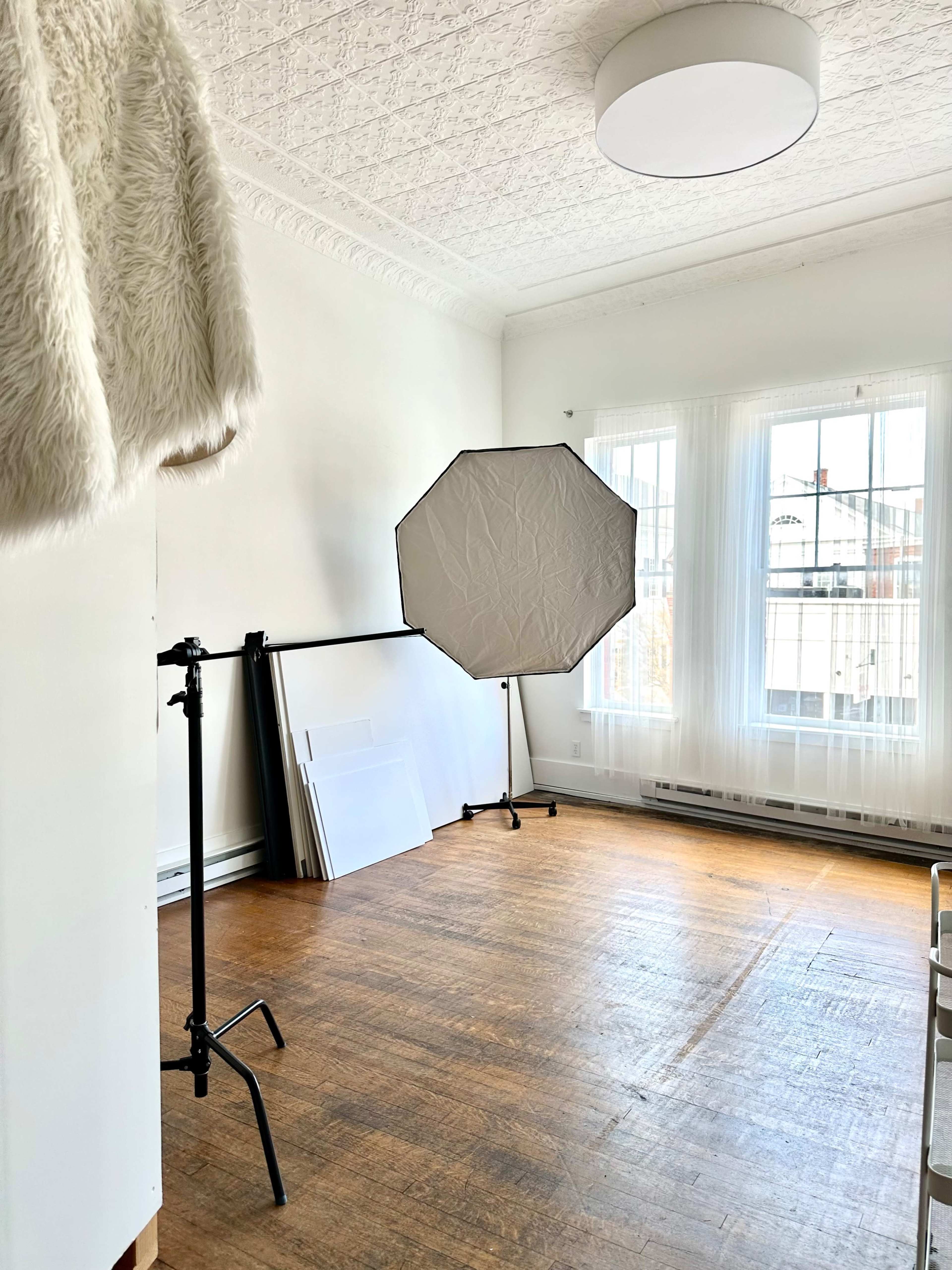 A sparsely furnished room with wooden floors, large windows, and a light stand holding an octagonal softbox.