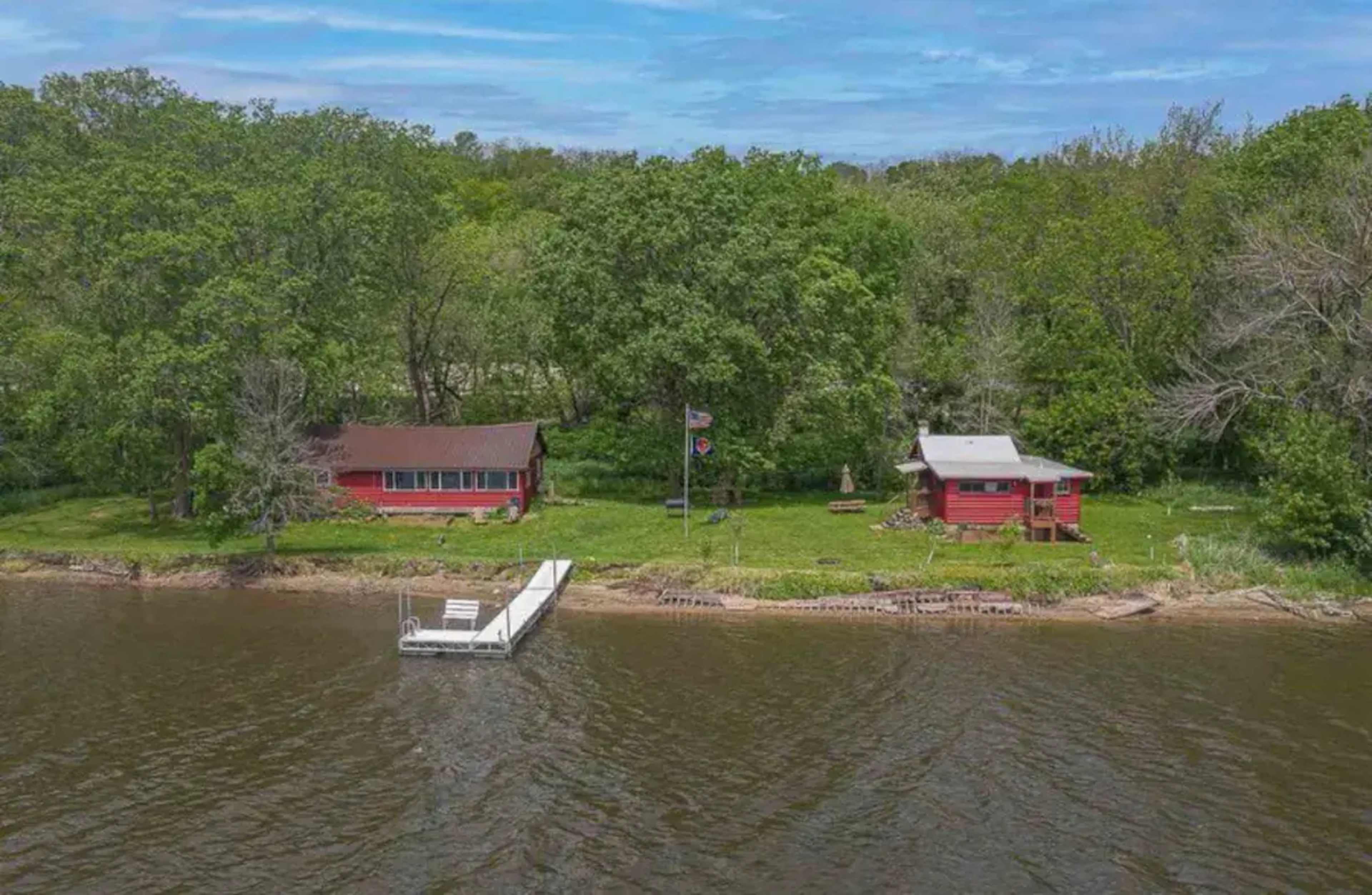 Two red cabins along a riverbank, surrounded by lush green trees, with a dock extending into the water.