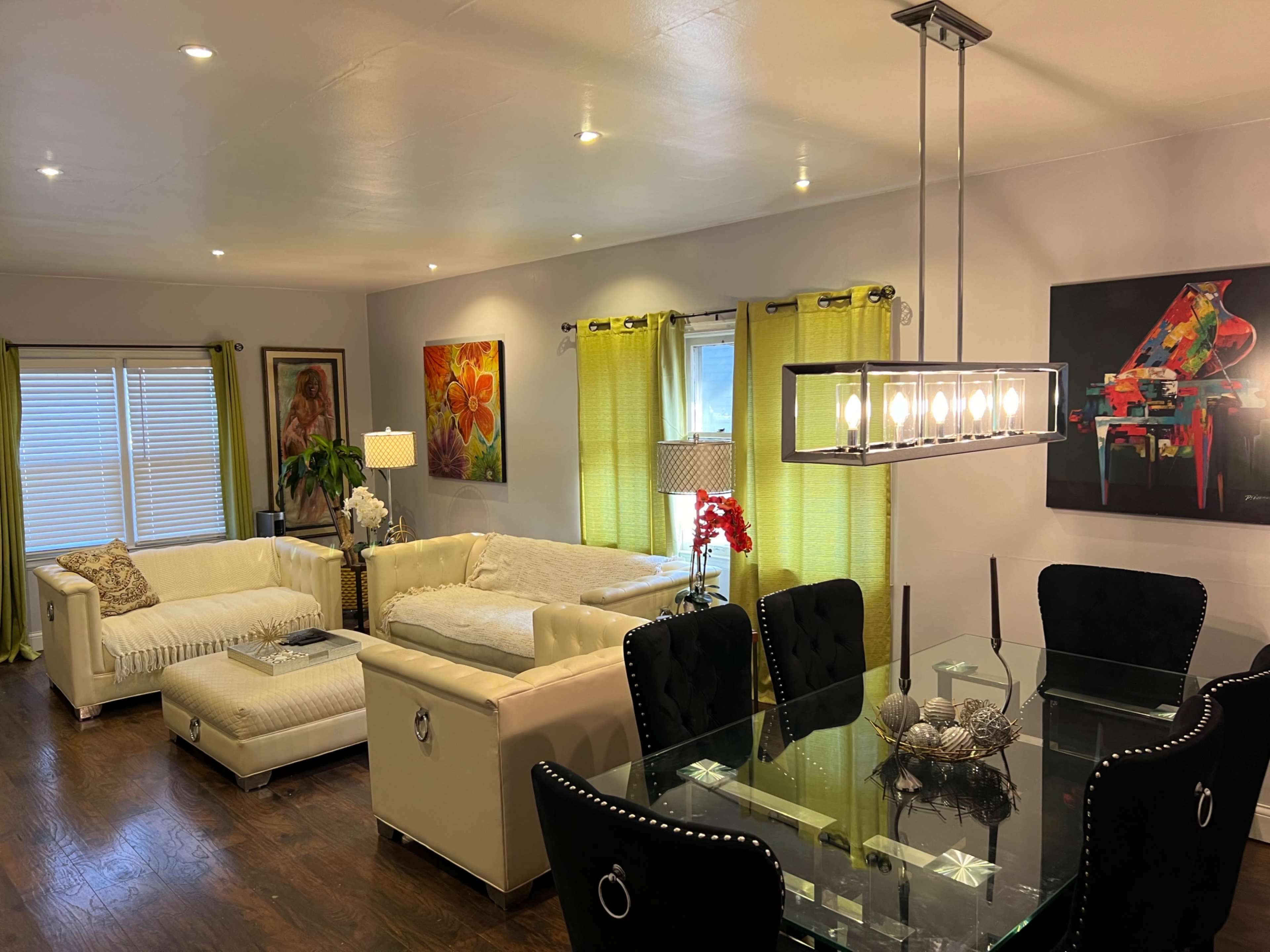 A modern living room and dining area with white furniture, colorful artwork on the walls, and large windows with green curtains.