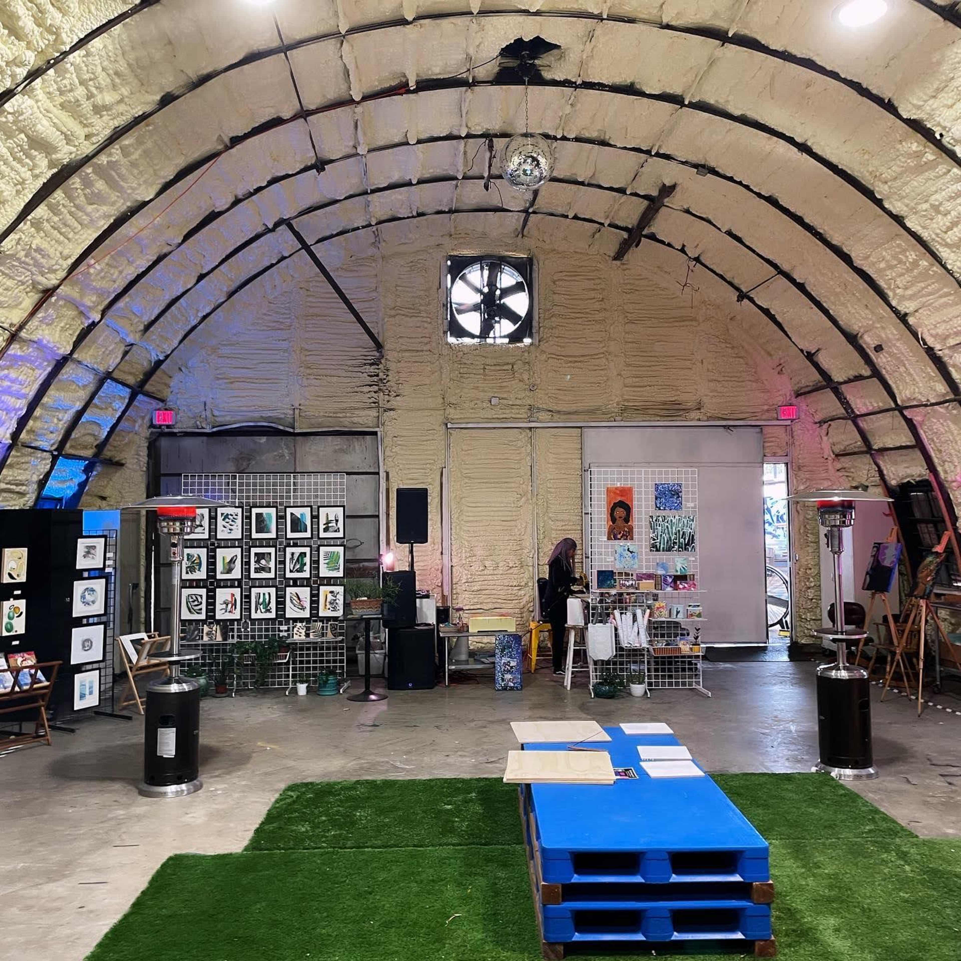 An art exhibition inside a large, curved structure with white textured walls, featuring various artworks displayed on grids and panels around a central area with a blue wooden platform and green turf.