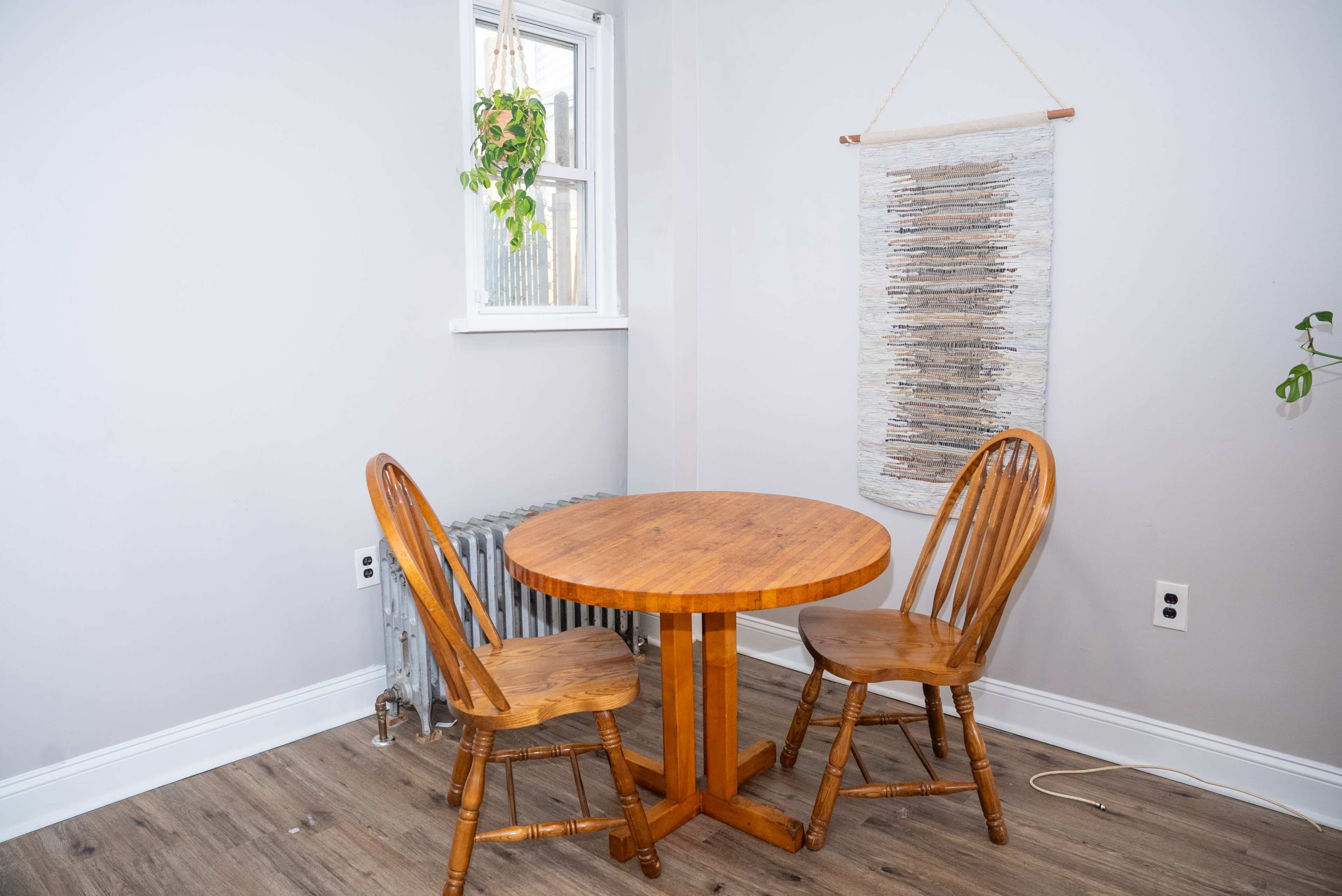 A small wooden table with two matching chairs is positioned against a light-colored wall, with a radiator and a hanging piece of artwork nearby.