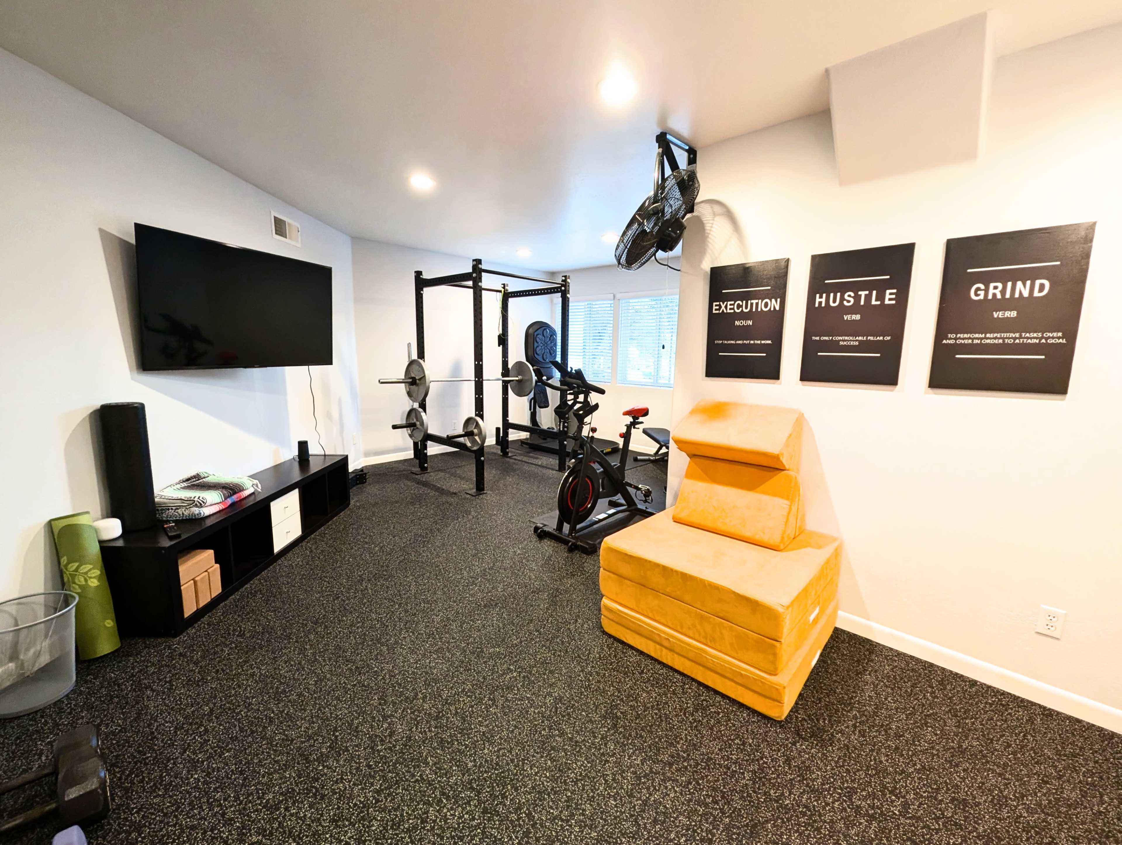 The image shows a modern home gym featuring a workout area with exercise equipment, a large TV, and motivational posters on the wall.