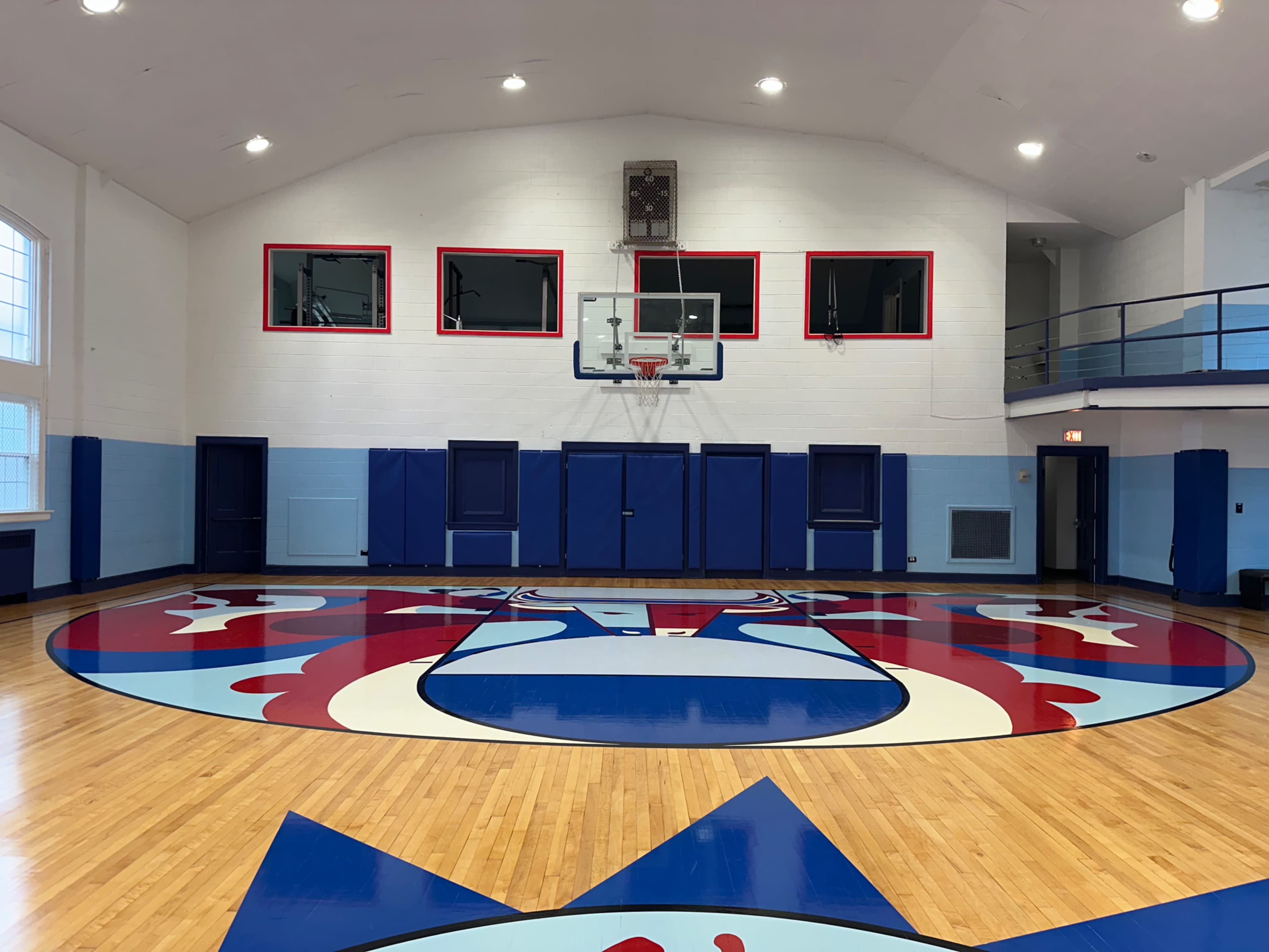 The image shows a basketball gymnasium with a hardwood floor featuring a painted logo at center court, surrounded by blue walls and large windows.