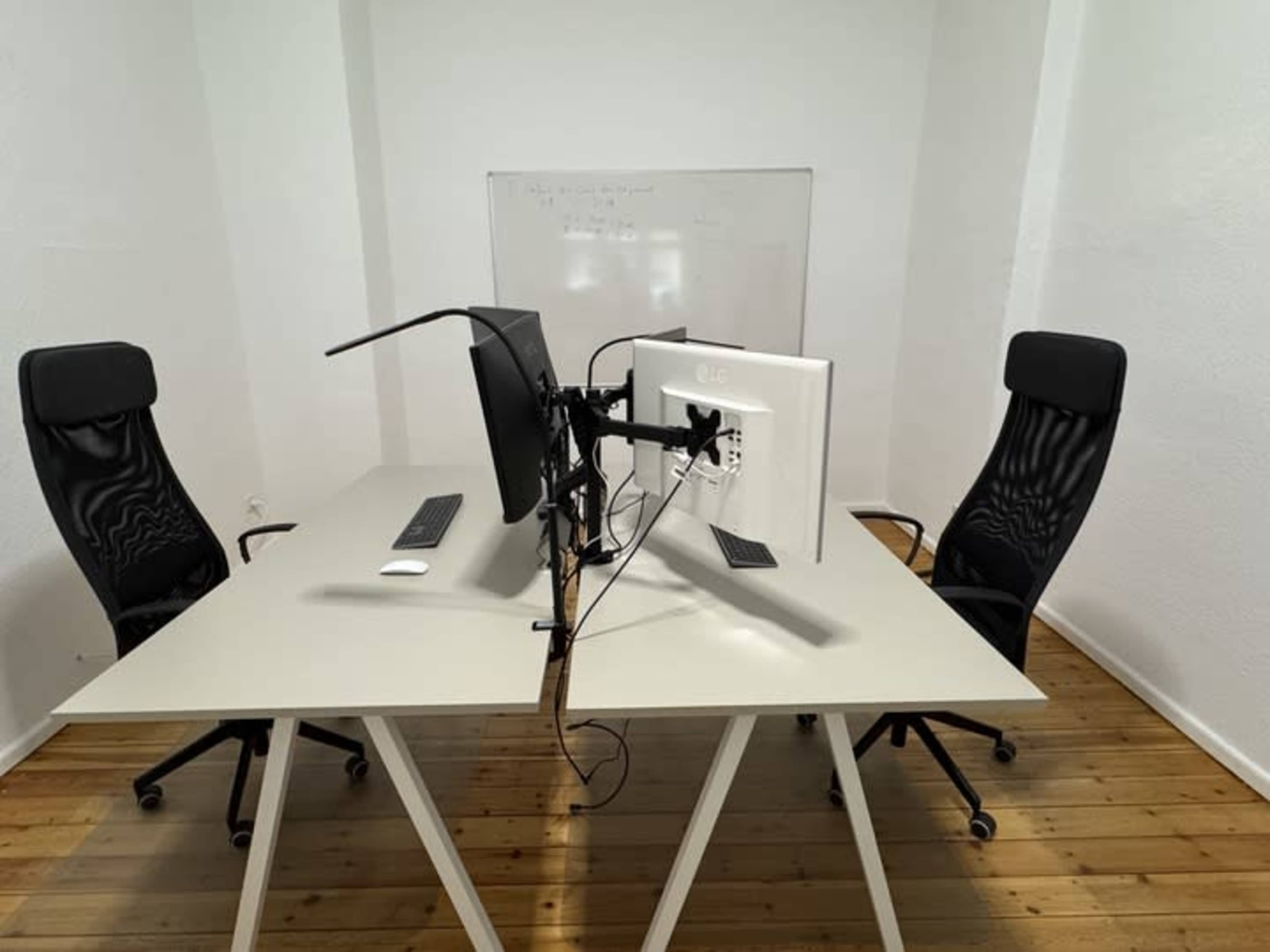 A minimalist office space features two modern chairs facing a shared desk with dual monitors and a whiteboard in the background.