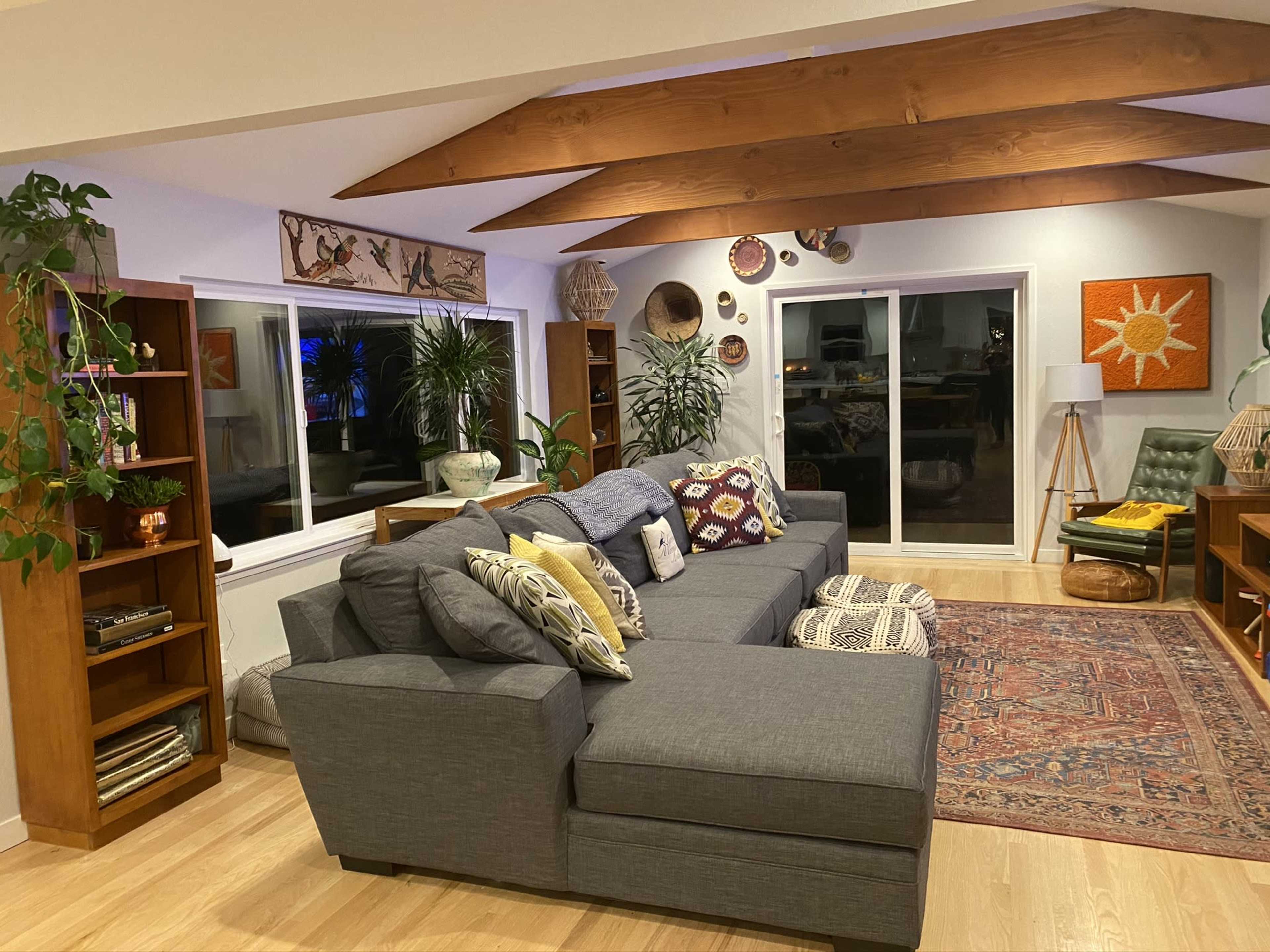 A living room features a large gray sectional sofa, wooden shelves filled with decor, and a colorful area rug beneath a ceiling with exposed beams.