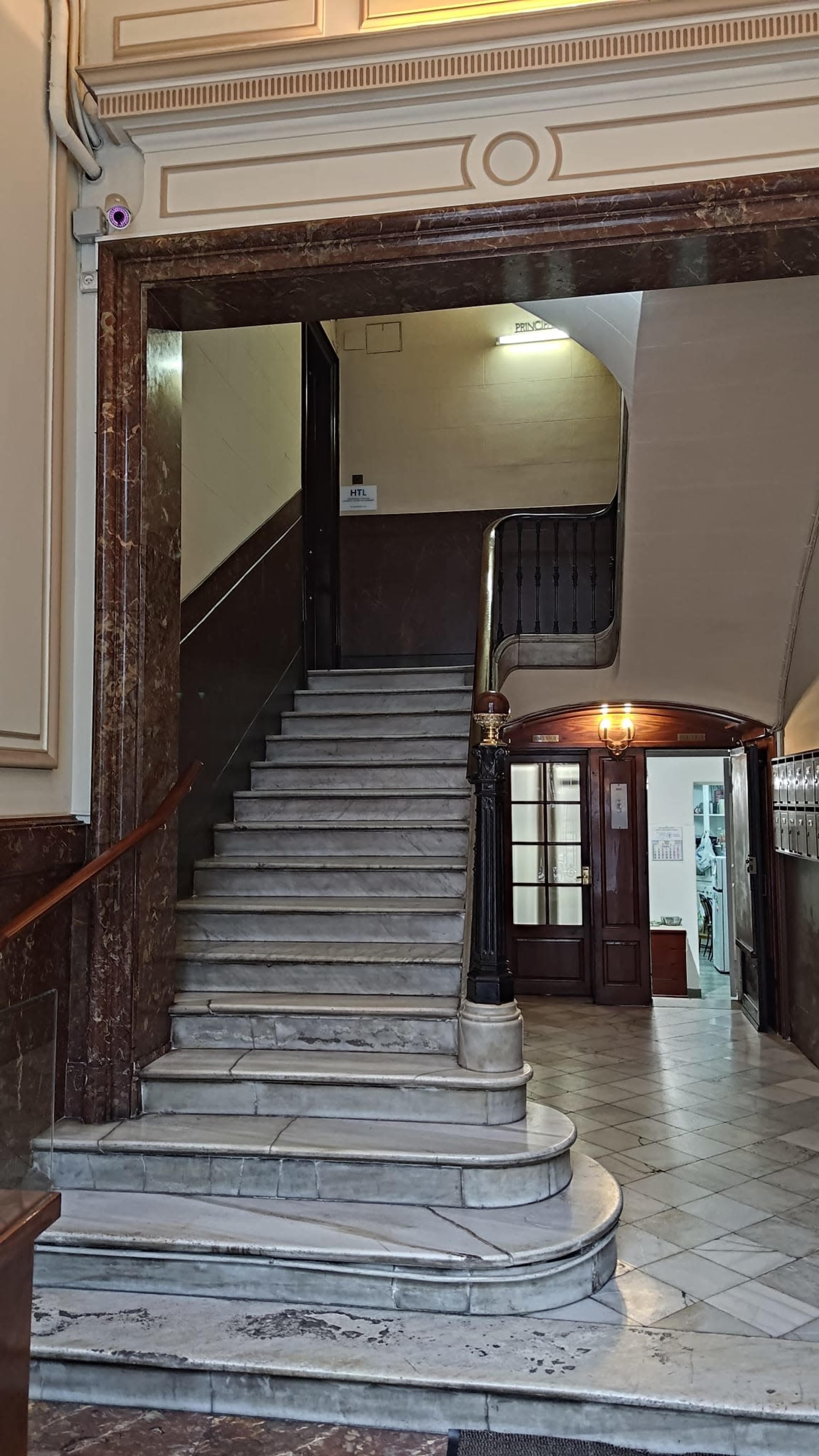 The image shows a marble staircase leading to an upper level in a building, with wooden railings and a tiled floor visible in the foyer area.