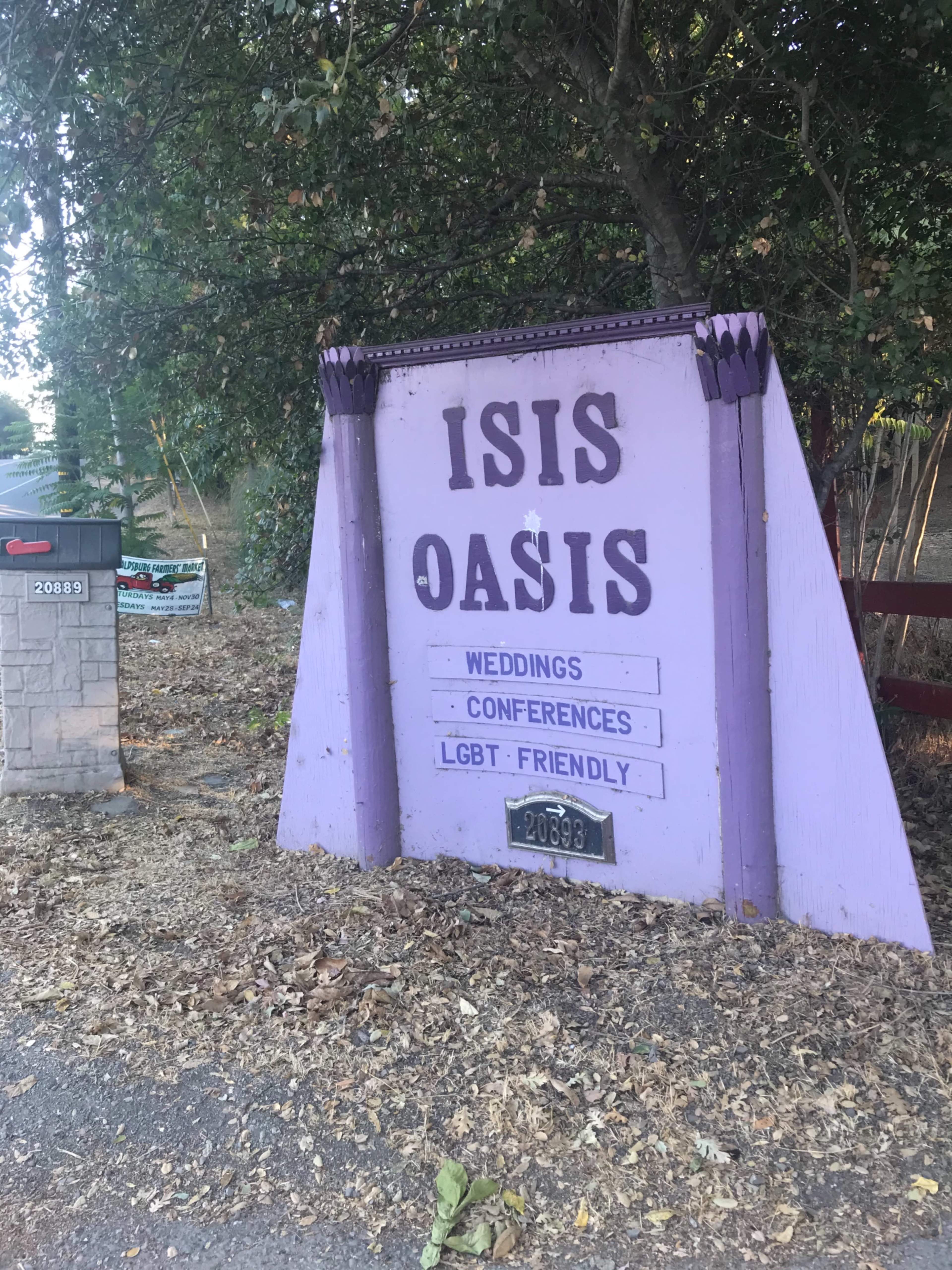 A purple sign for "Isis Oasis" indicating it is a venue for weddings, conferences, and is LGBT friendly, situated near a tree line.