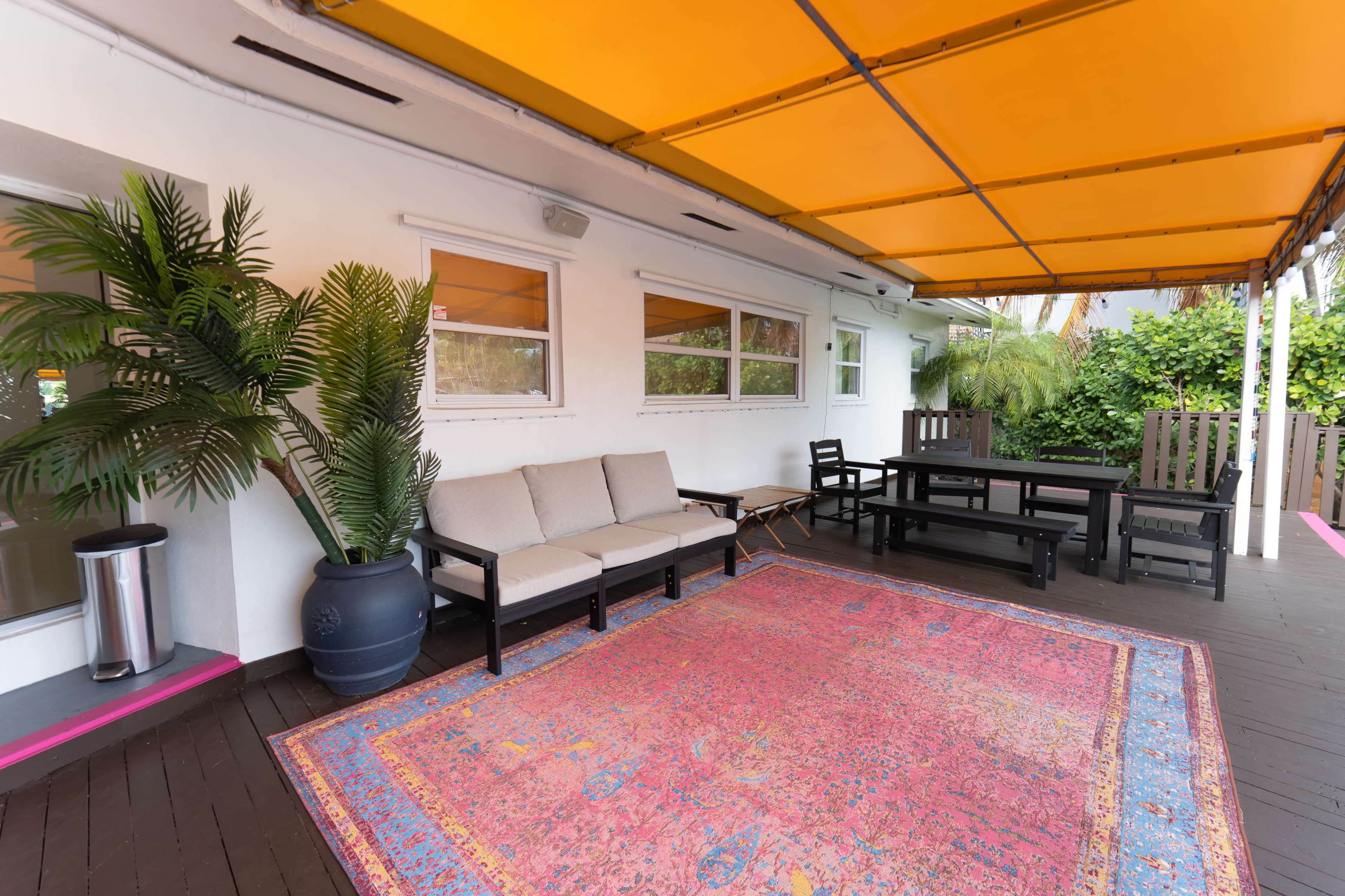The image shows a covered outdoor space featuring a large patterned rug, a sofa, a dining table, and potted plants against a backdrop of white walls and a bright yellow awning.