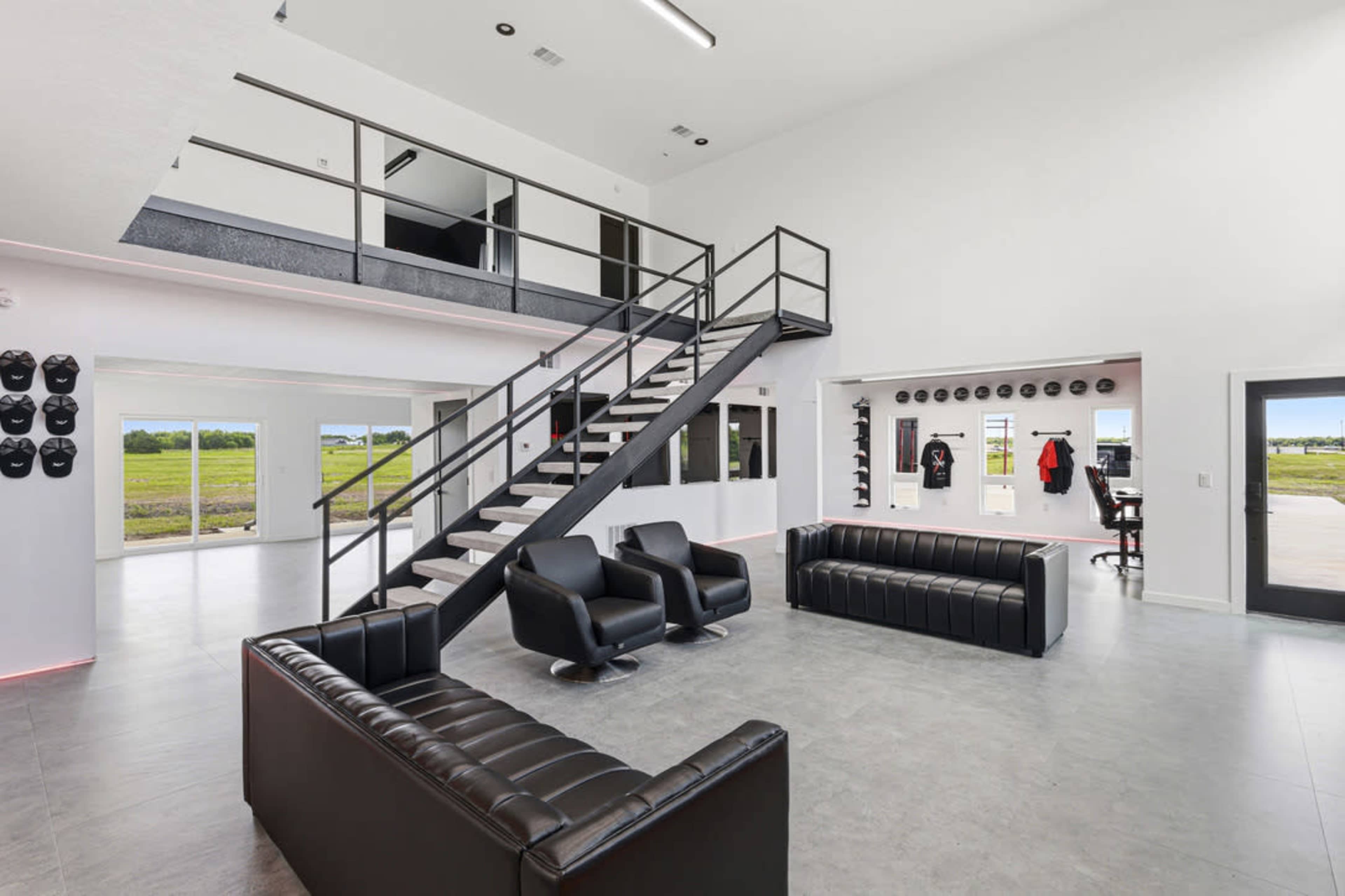 The image shows a modern interior space with a double-height ceiling, a staircase leading to a second level, and a seating area featuring black leather couches.