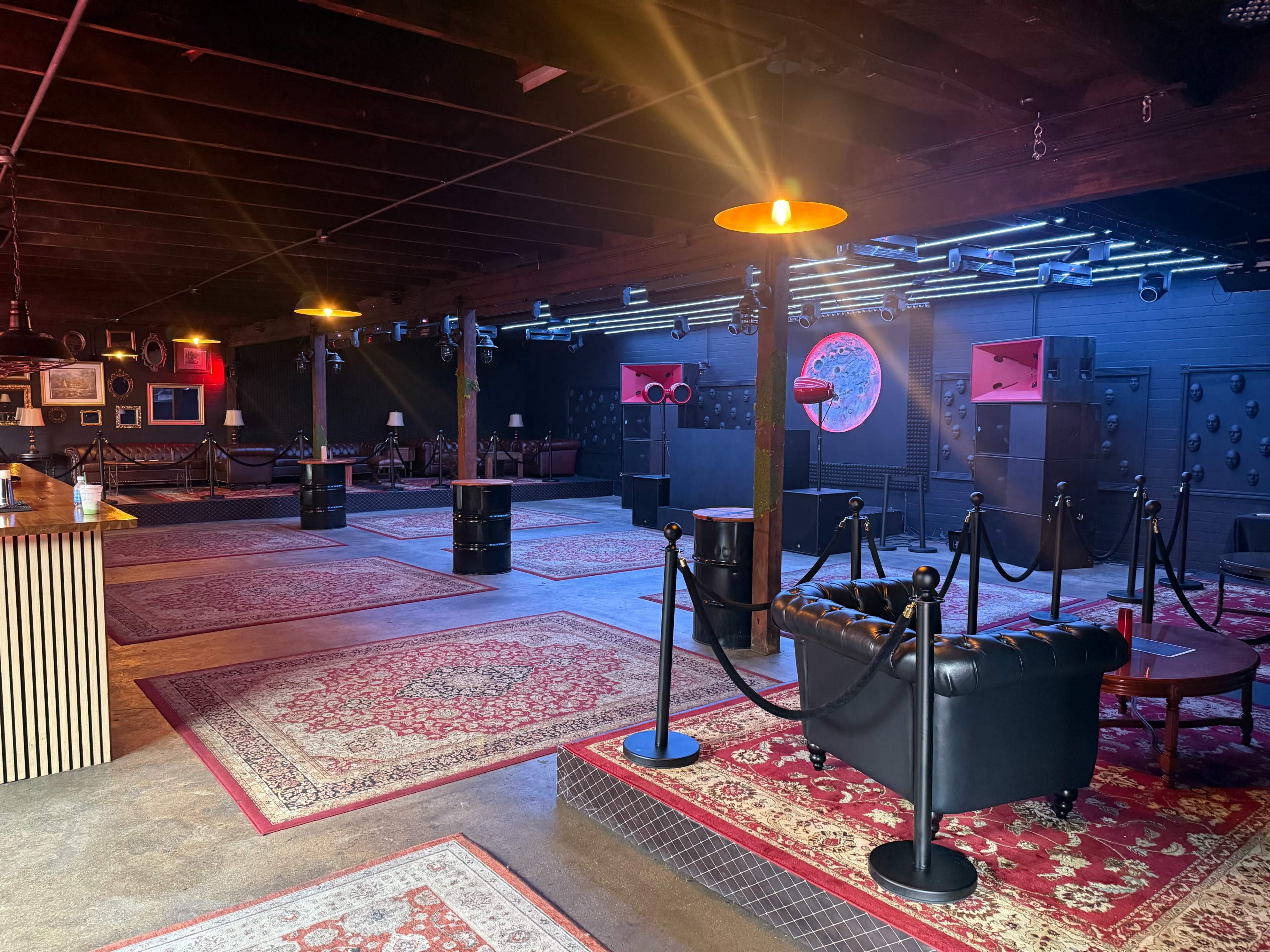 The image shows an empty music venue interior with patterned rugs, stage speakers, and a seating area featuring a black couch and a bar.