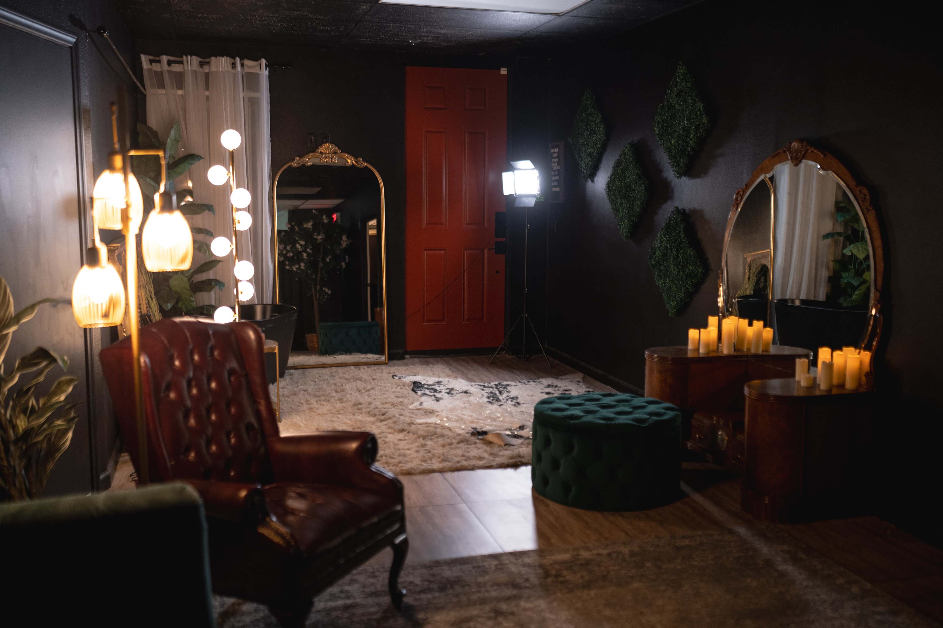 A dimly lit room with two ornate mirrors, a vintage armchair, a green upholstered stool, and candles arranged on a table, along with decorative plants and a red door in the background.