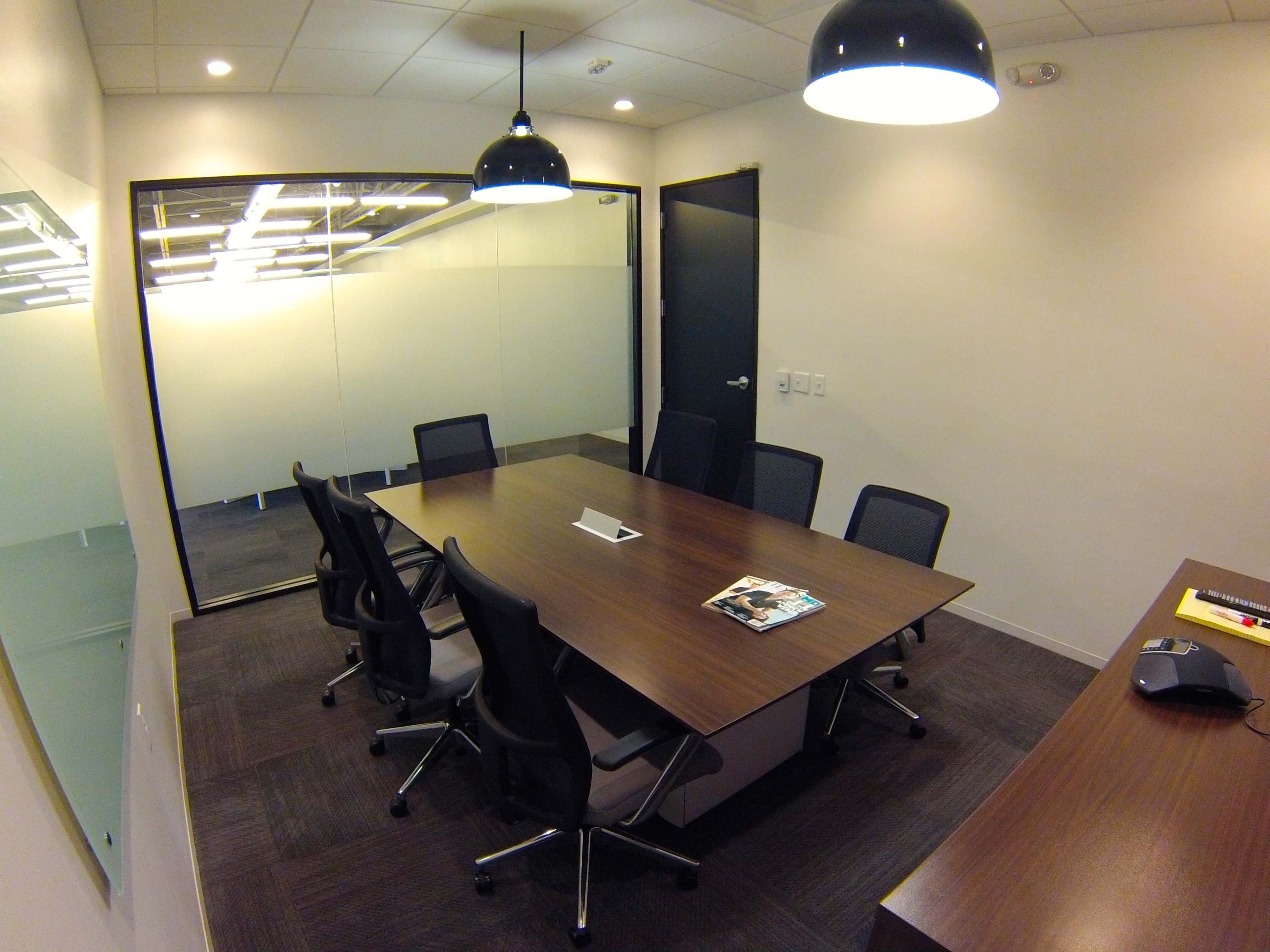 A conference room with a large table surrounded by black chairs, featuring a glass wall and modern lighting fixtures.
