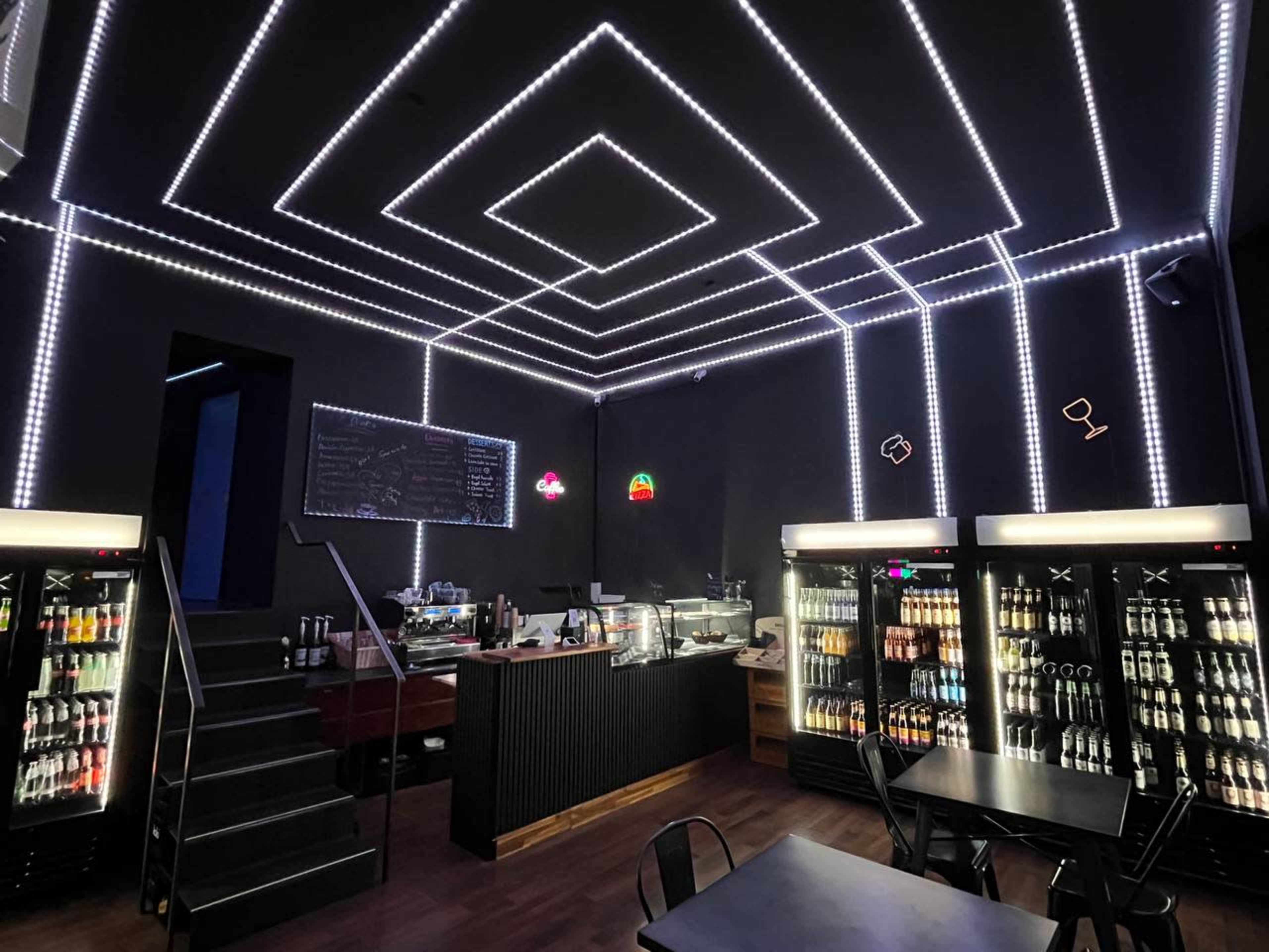 The image shows a modern café with neon lighting, a beverage display, and a staircase leading to another level.