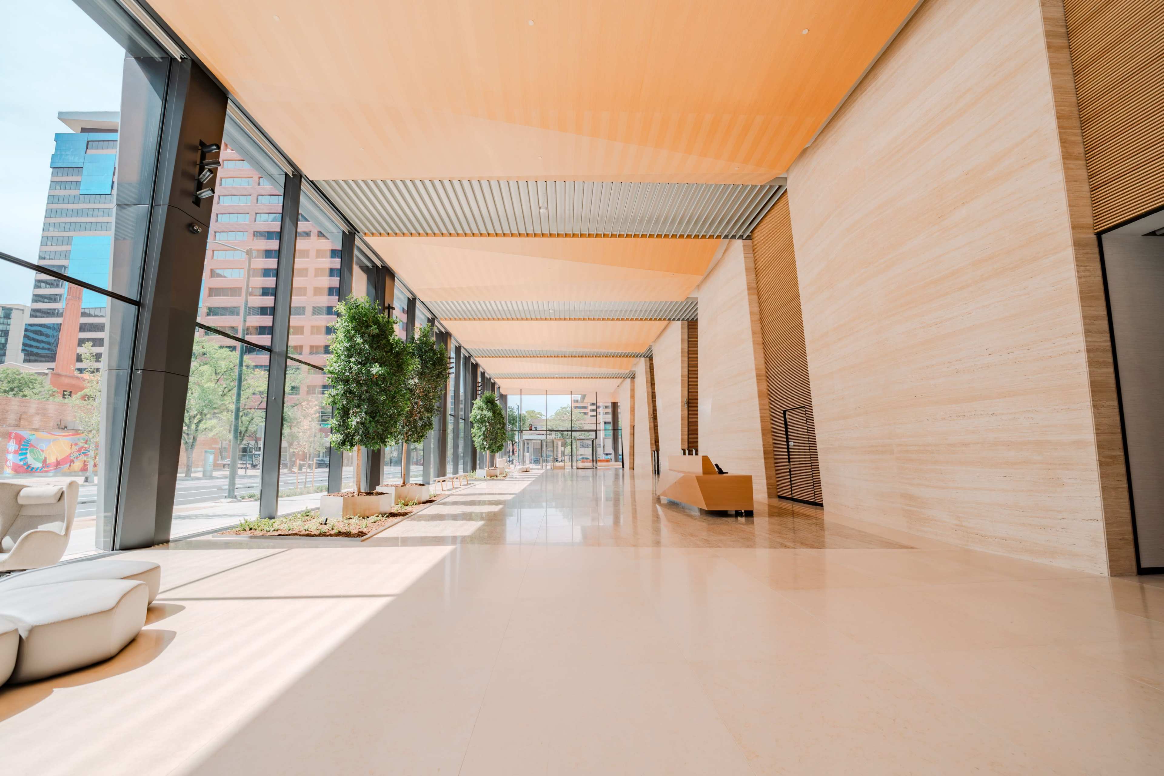 The image shows a modern, spacious lobby with large windows, light wooden ceilings, and potted trees lining the floor.