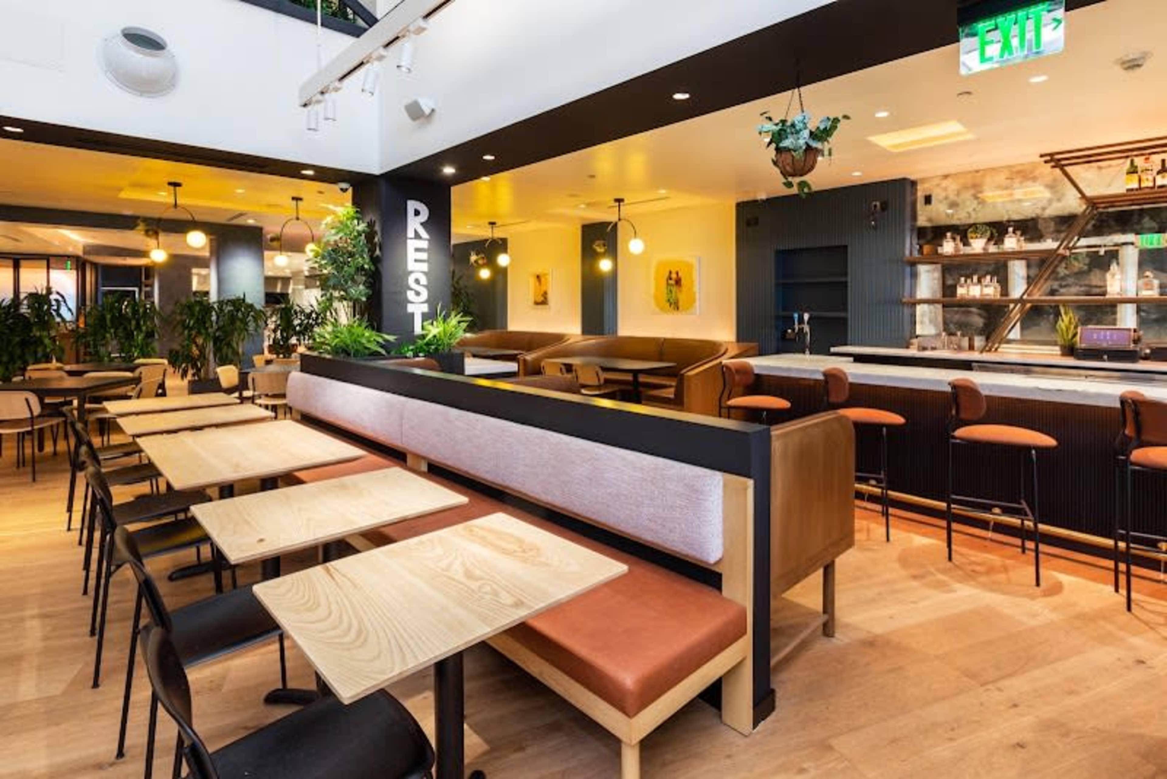 The image shows a modern restaurant interior with wooden tables, brown seating, and plants, featuring a bar area in the background.