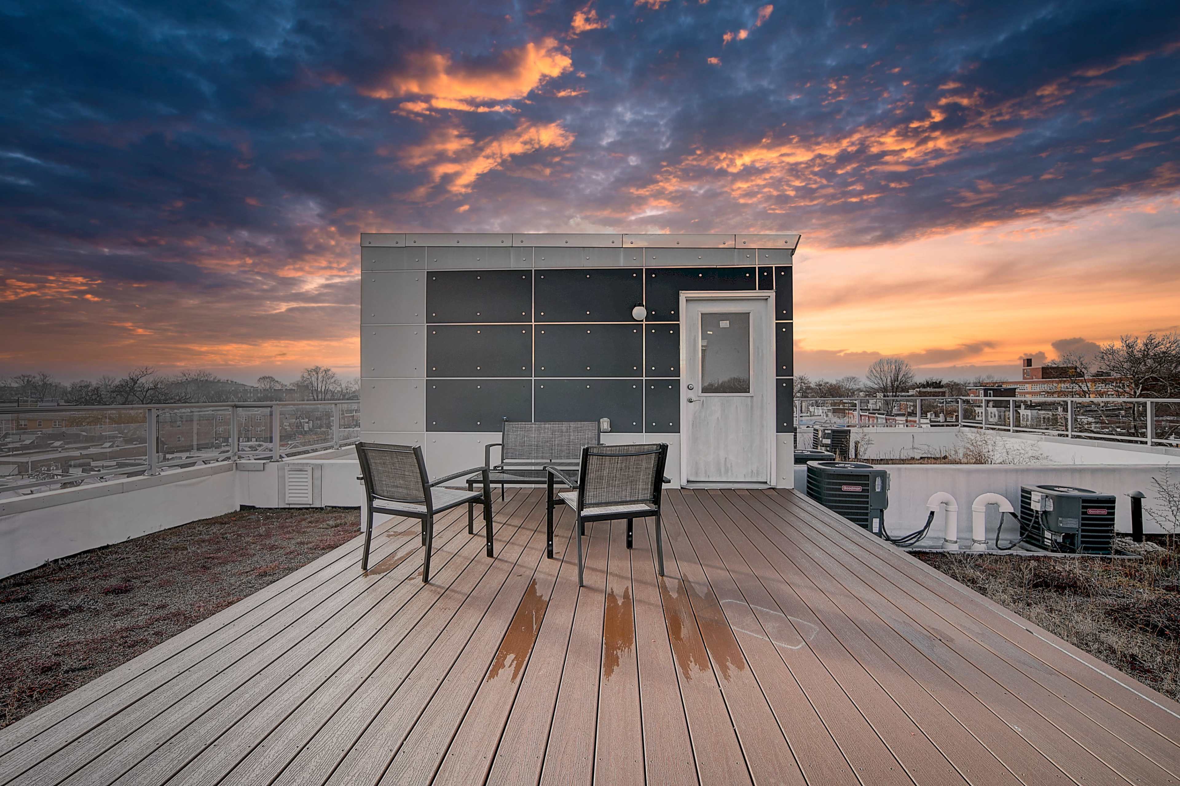 A rooftop deck features four chairs arranged around a small table, with a modern building and a colorful sunset in the background.