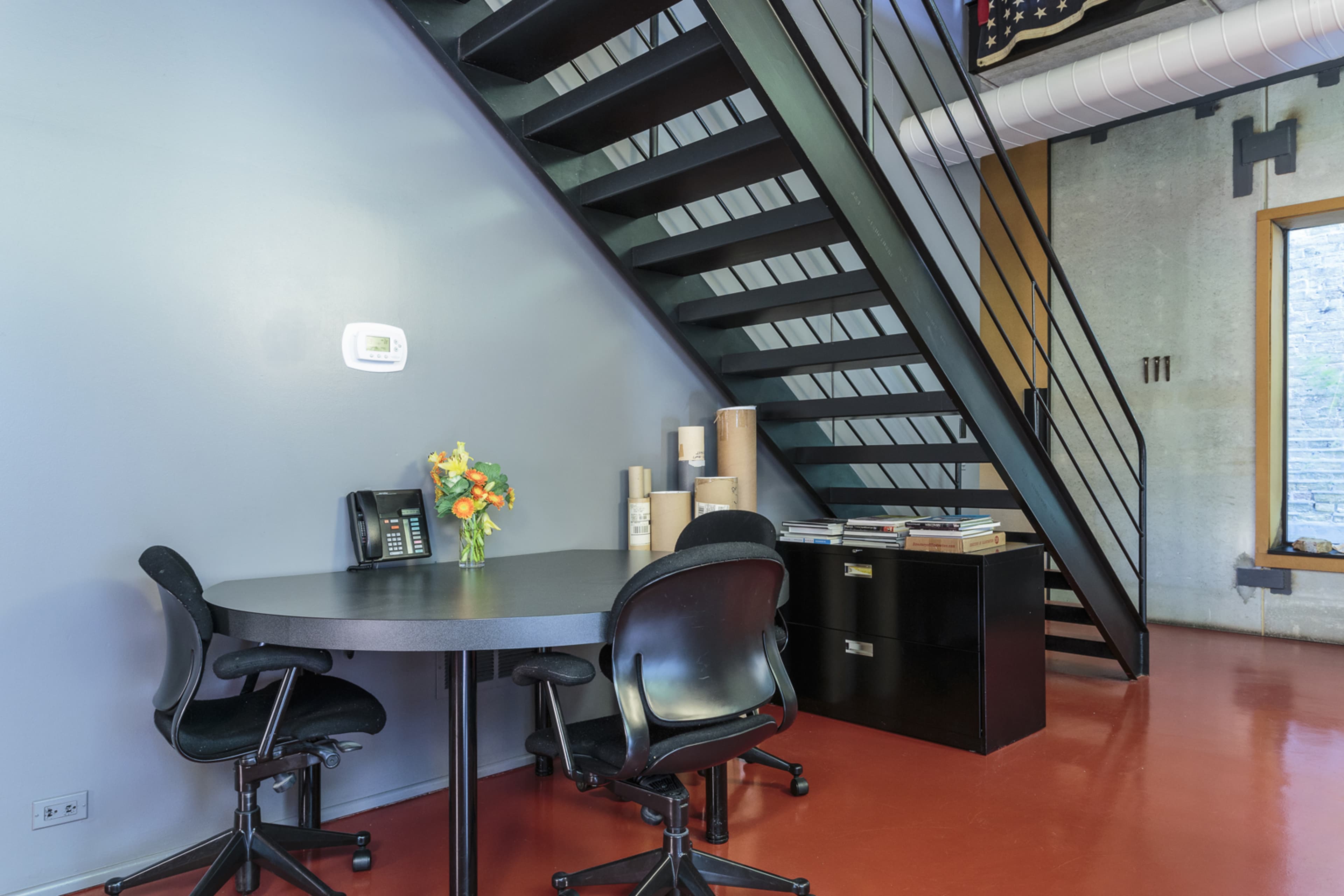 A modern office space with a round table, two black chairs, a telephone, a small bouquet of flowers, a filing cabinet, and stairs leading to an upper level.