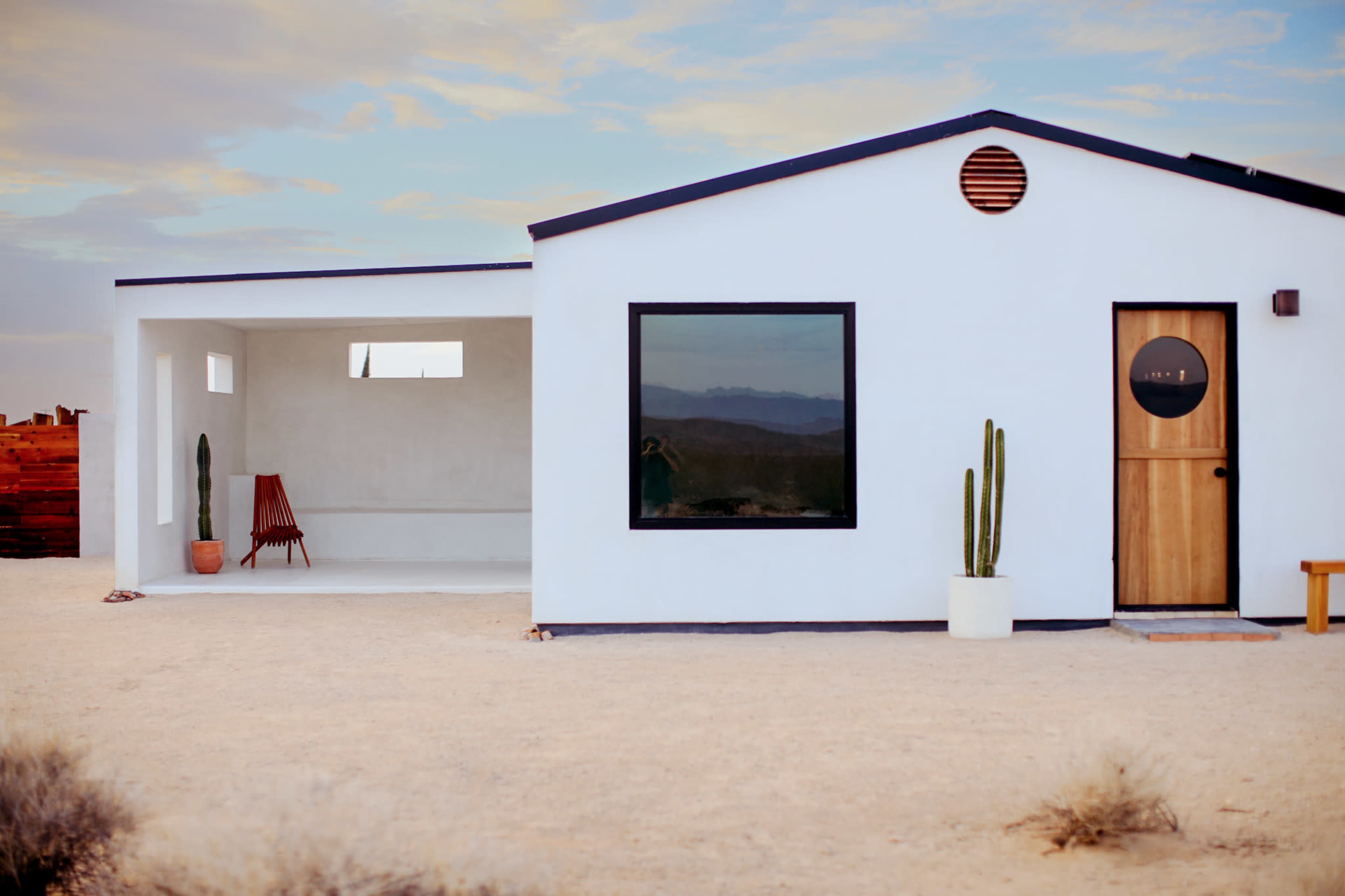 A white adobe-style house with a wooden door and large windows stands in a desert landscape, accompanied by a red chair and a potted cactus.