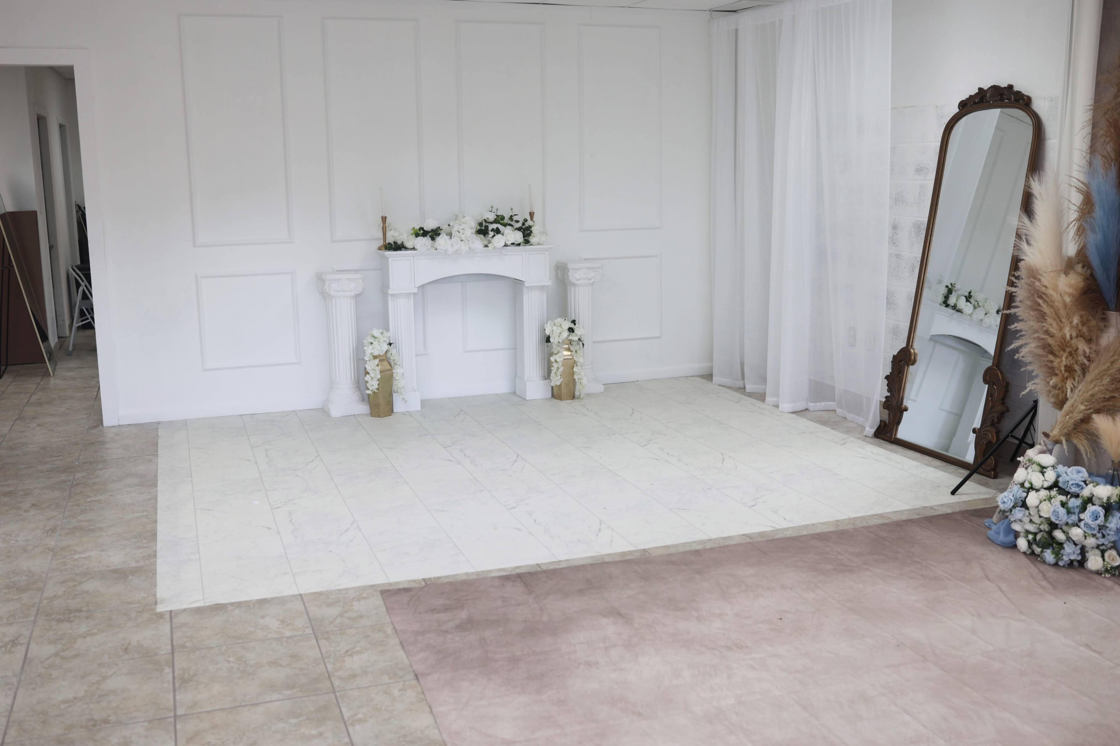 The image shows a minimalistic interior space featuring a white fireplace facade with decorative flowers, a large mirror, and light-colored flooring.