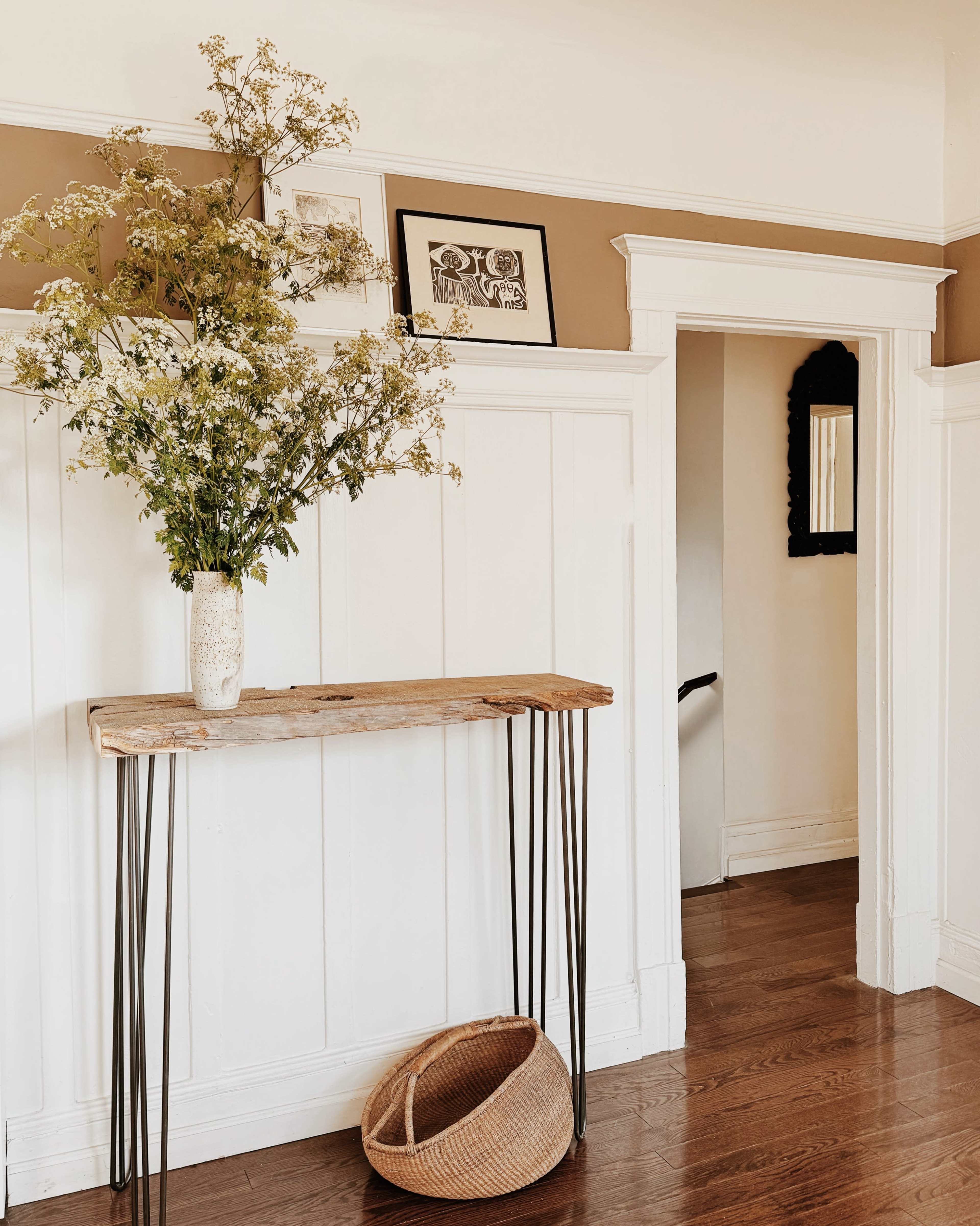 A wooden console table with metal legs holds a vase of flowers against a white-paneled wall, with a doorway leading to another room visible in the background.