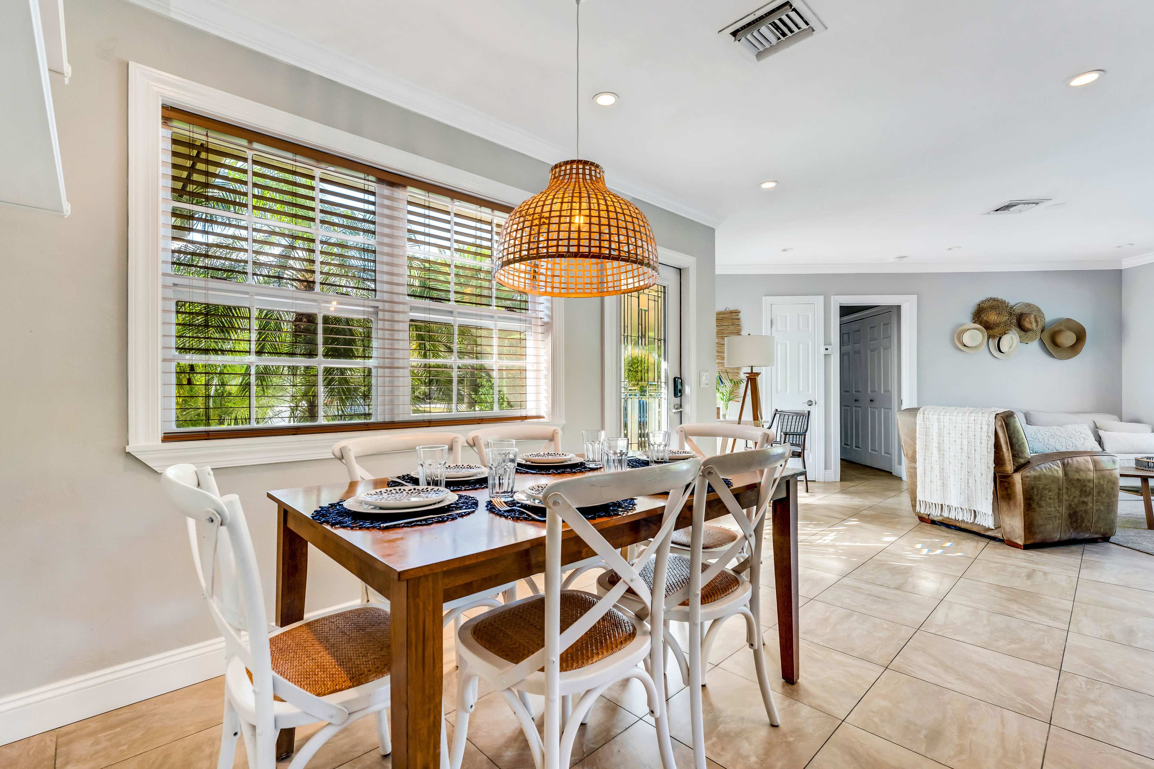 A dining area features a wooden table set for four, with a woven pendant light above and a view of large windows letting in natural light.
