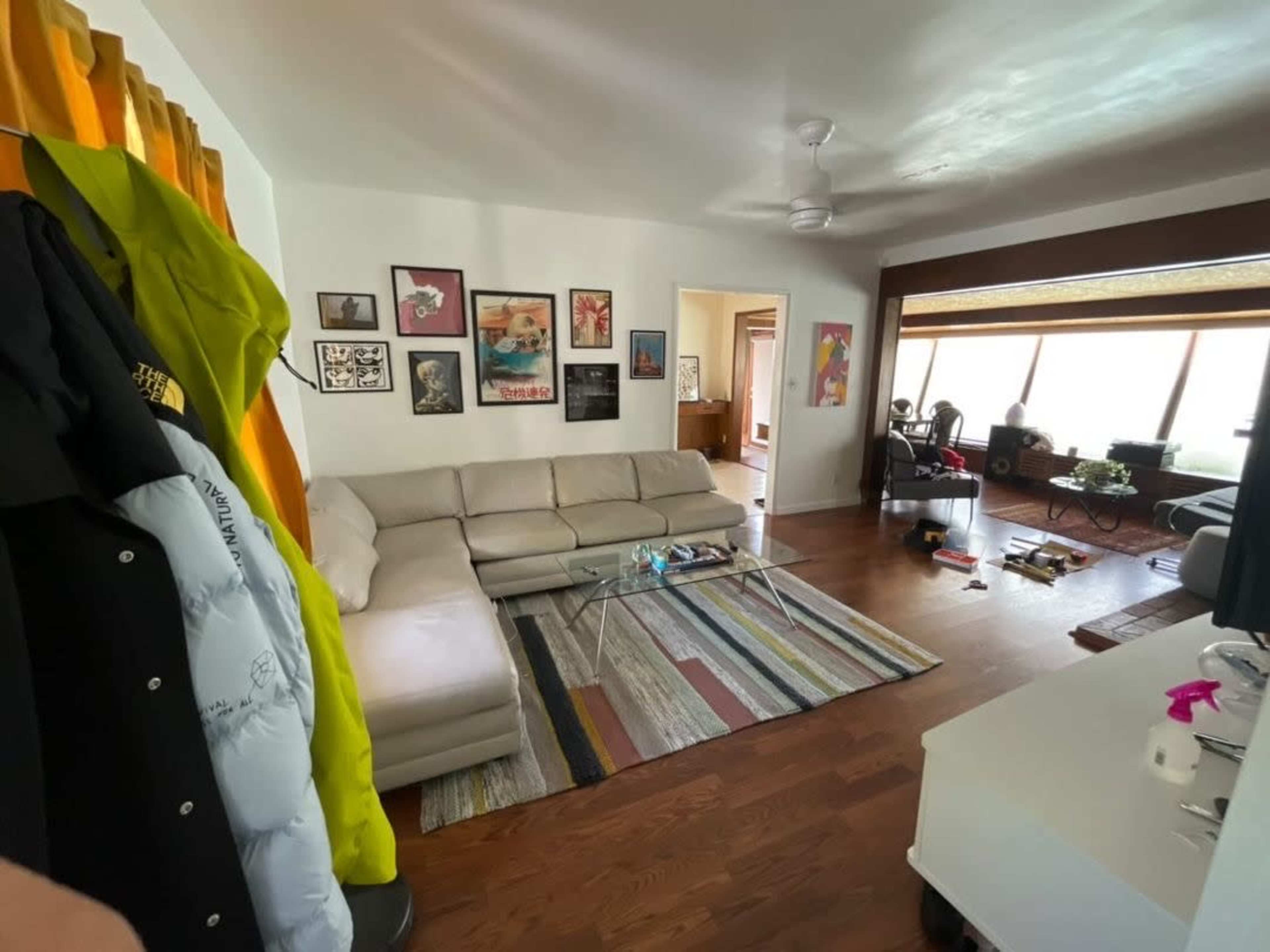 A living room with a sectional sofa, a glass coffee table on a striped rug, framed artwork on the walls, and various items scattered on the floor.
