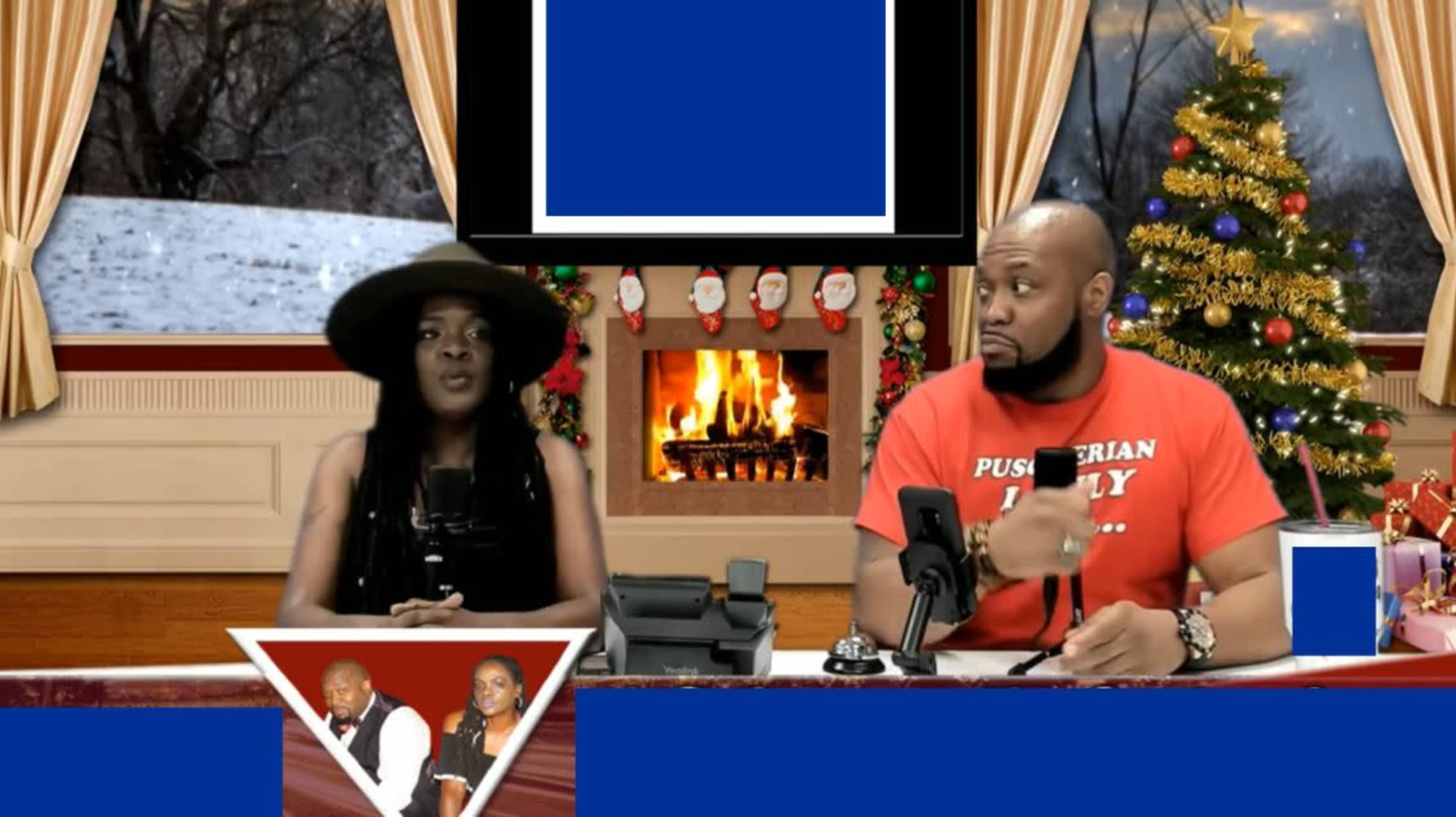 A woman and a man sit at a desk in a studio decorated for the holidays, with a fireplace and snowy scene visible through the windows behind them.