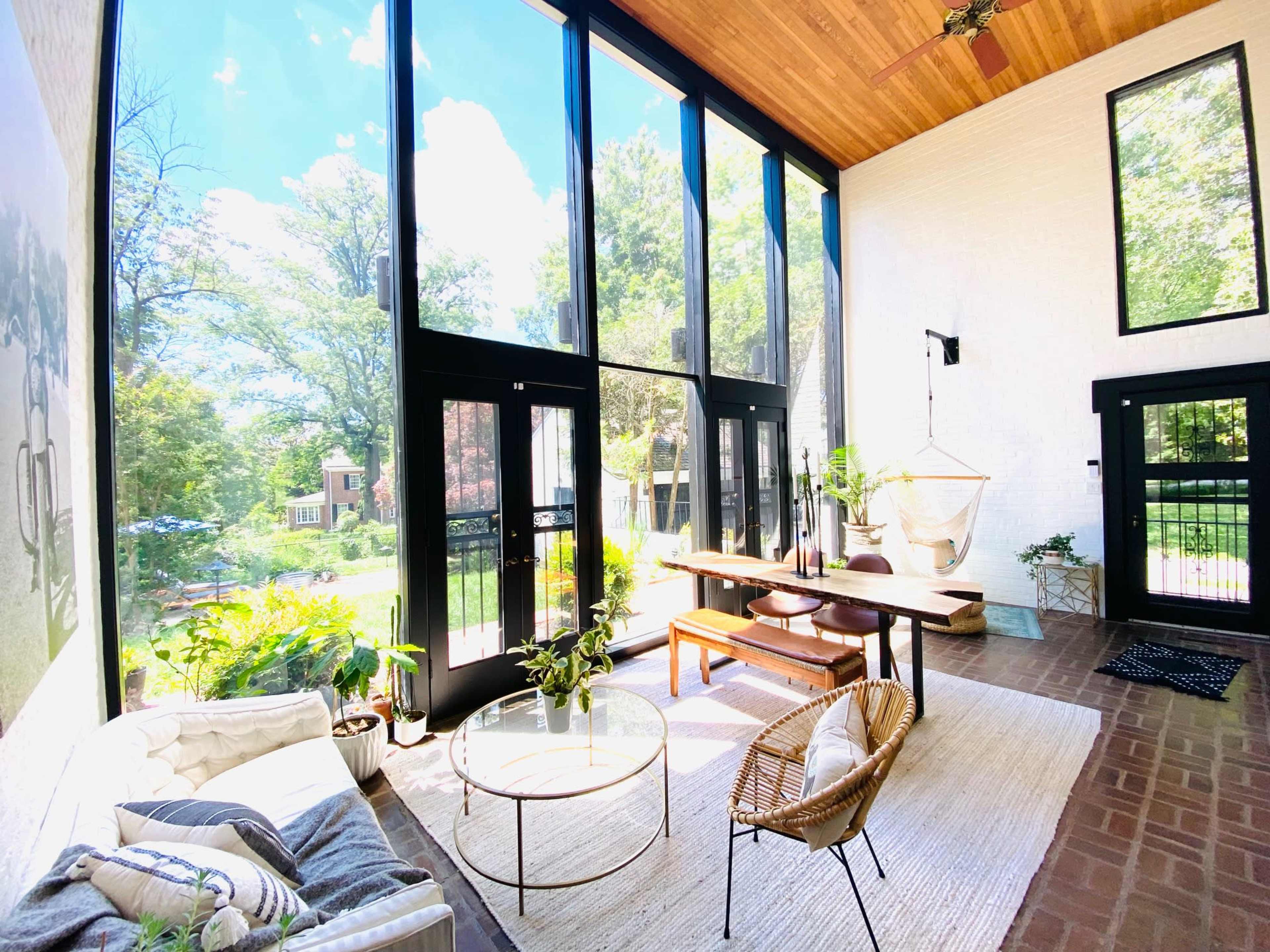 A bright and airy sunroom features large glass windows, a wooden ceiling, a dining table, and various plants.