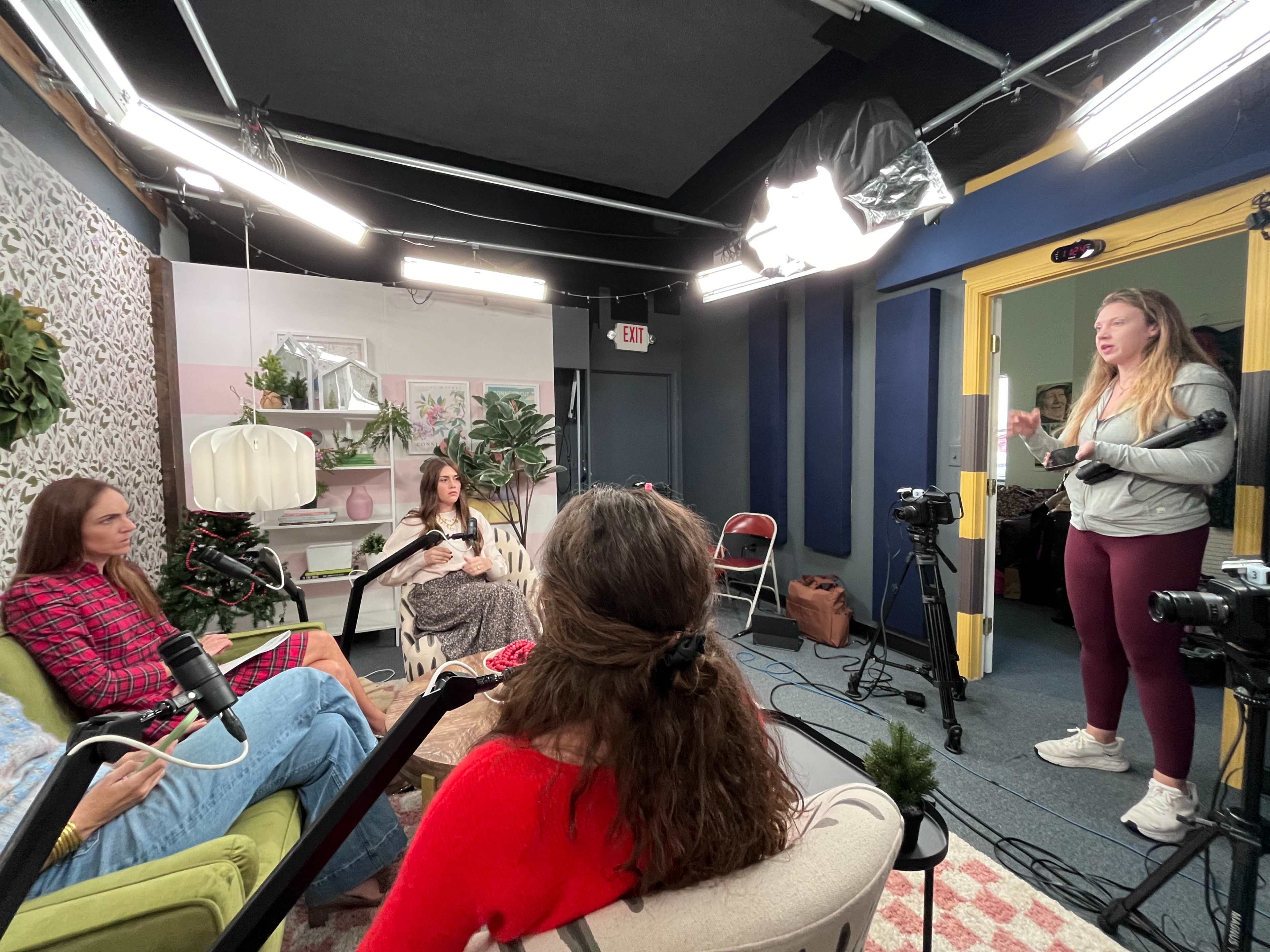 A group of four women is engaged in conversation in a well-lit studio set, featuring a green couch, potted plants, and video cameras.