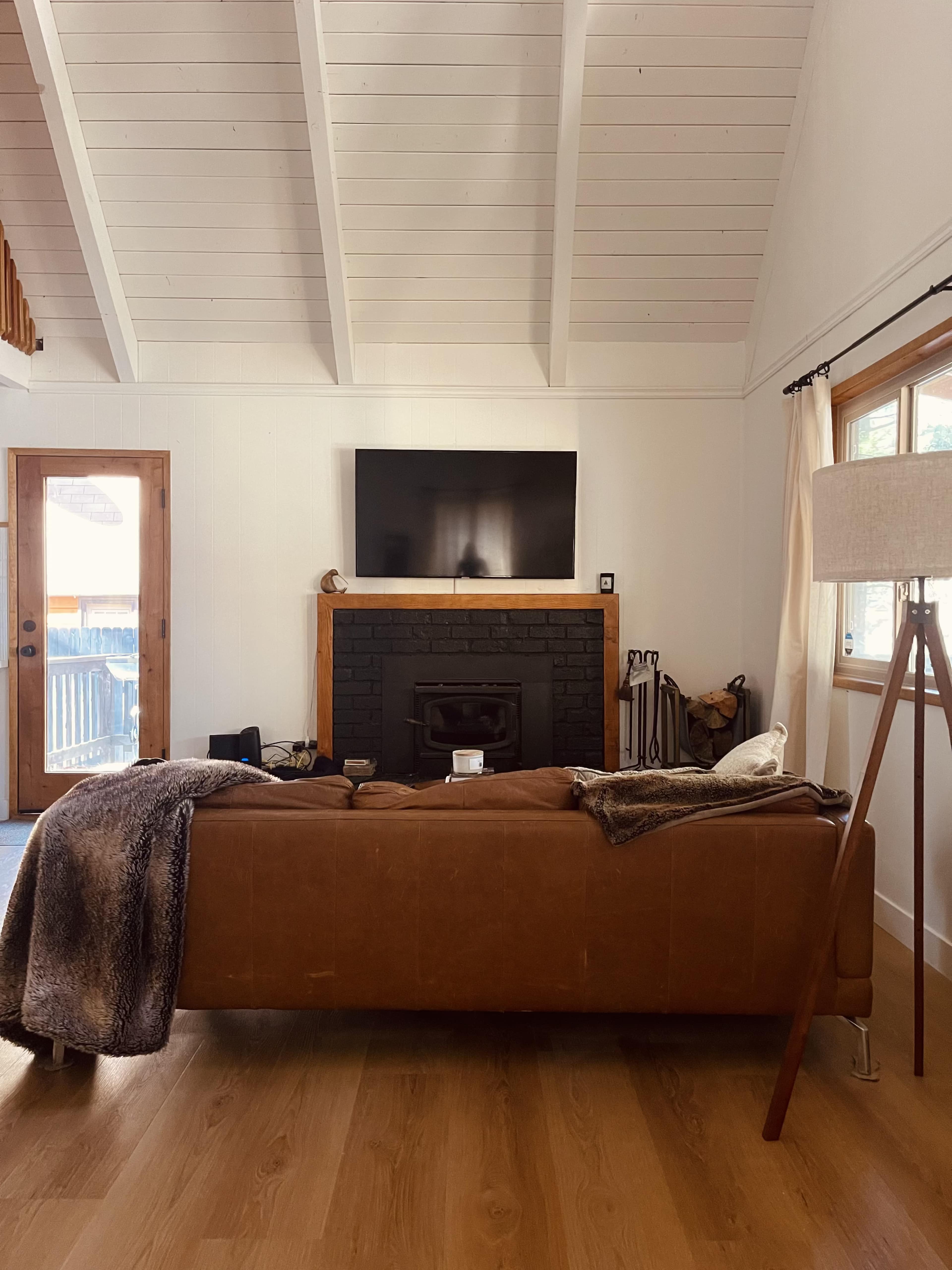 The image shows a cozy living room with a brown leather sofa facing a black fireplace and a television mounted on the wall.