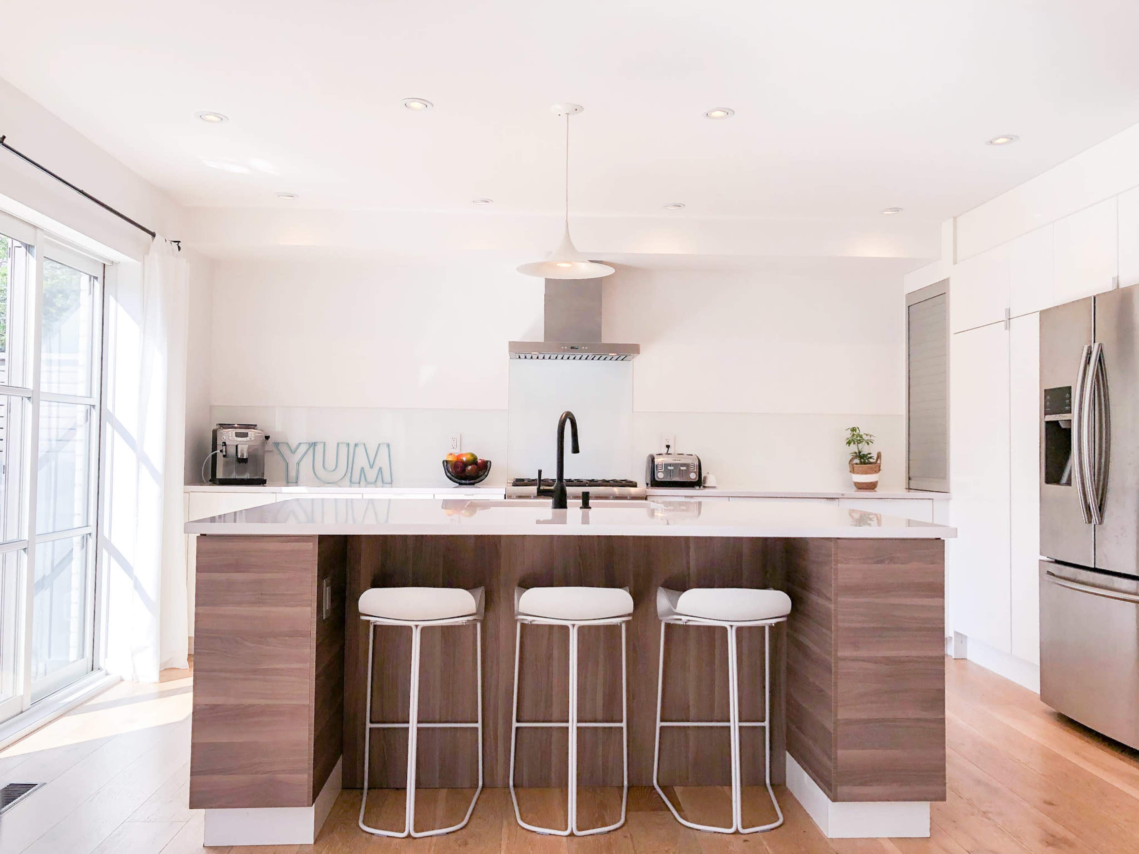 A modern kitchen features a central island with three bar stools, stainless steel appliances, and a sleek white countertop.