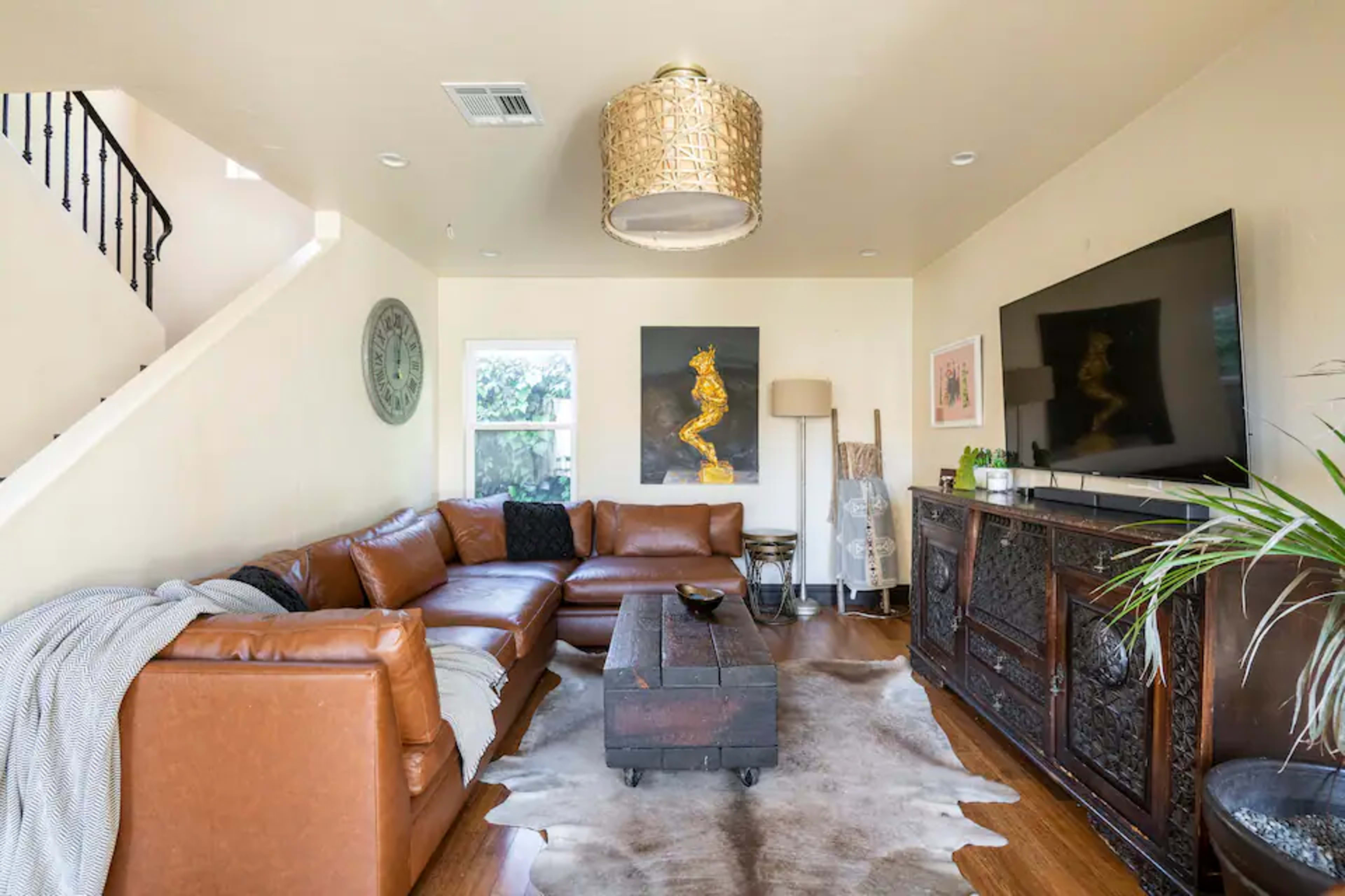 A spacious living room features a brown leather sectional sofa, a wooden coffee table, and a mounted television, with decorative artwork and a staircase in the background.