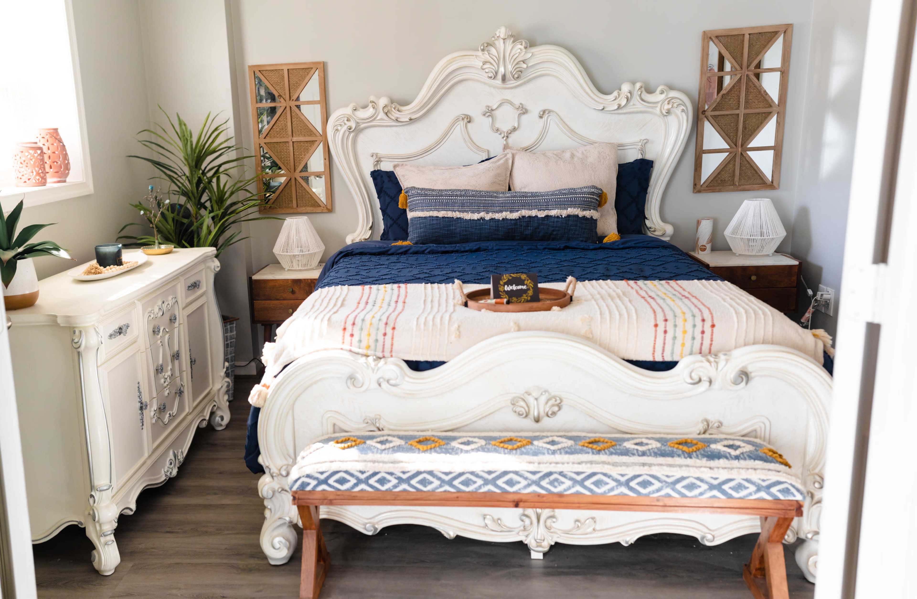 A decorated bedroom featuring a large ornate bed with blue bedding, a wooden bench at the foot, and two decorative mirrors on the walls.