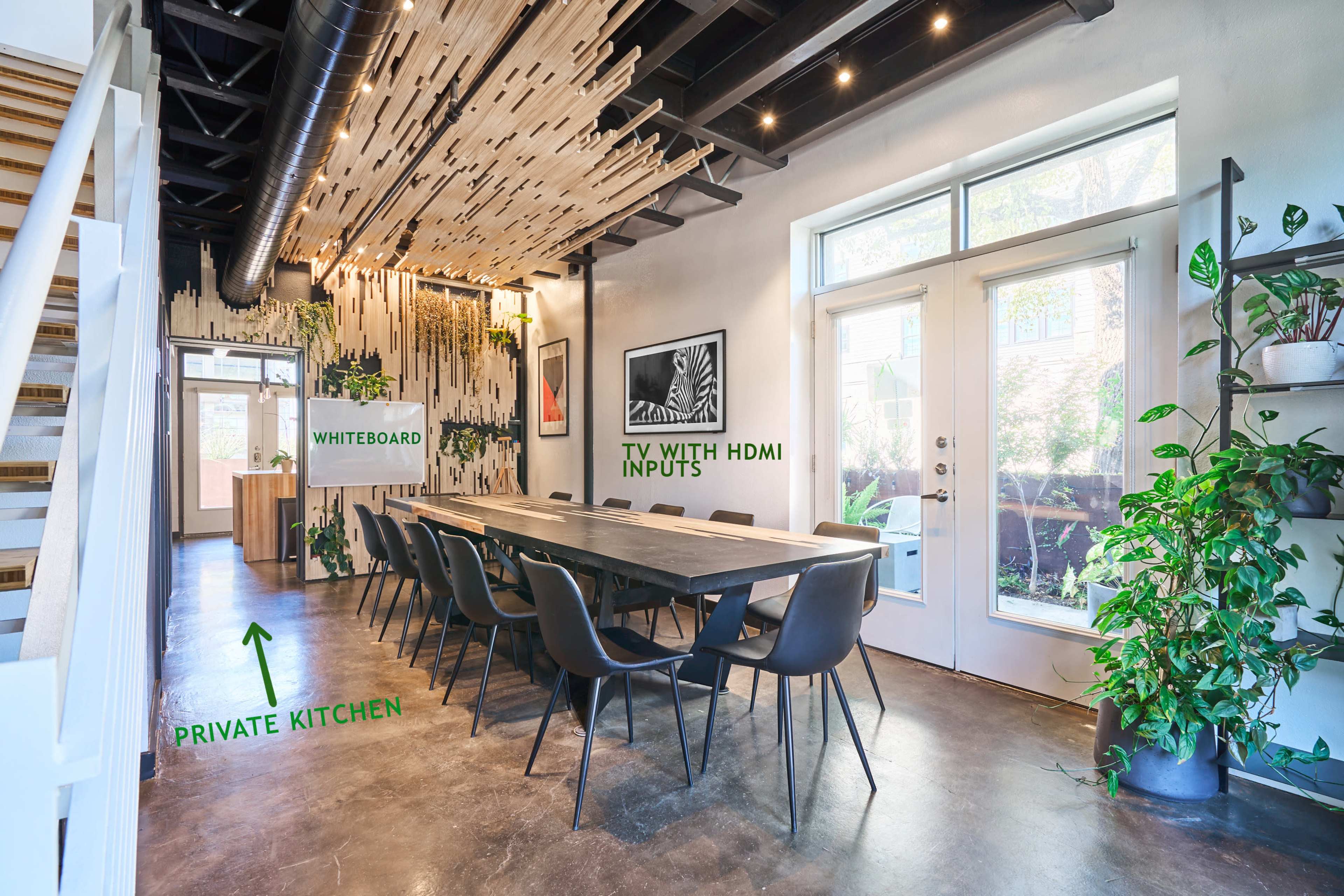 A modern meeting space featuring a long wooden table surrounded by black chairs, a whiteboard on the wall, and a TV with HDMI inputs, alongside a private kitchen area.