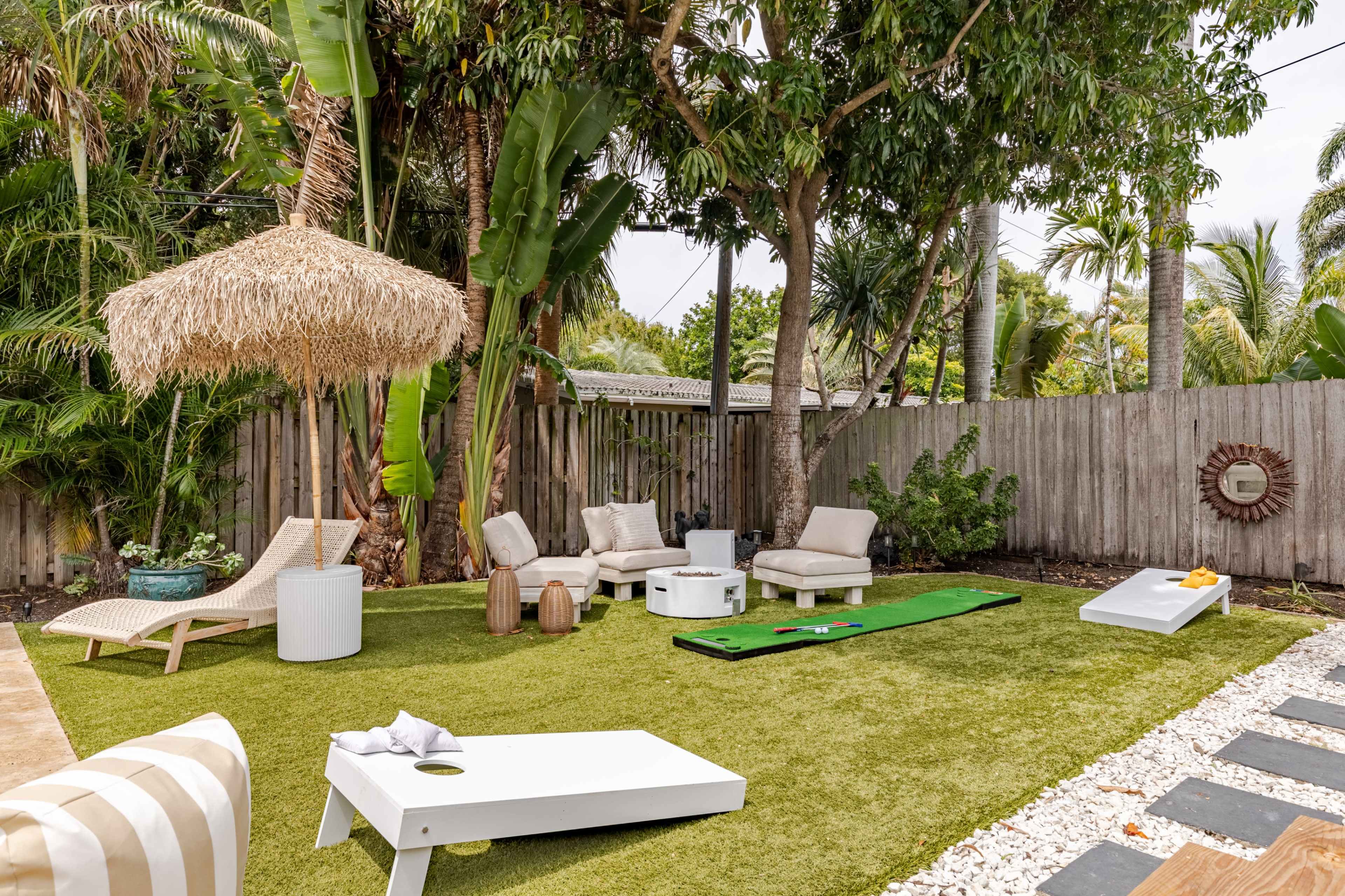 The image shows a landscaped backyard featuring a putting green, lounge chairs, a thatched umbrella, and a cornhole game on a lawn surrounded by tropical plants.