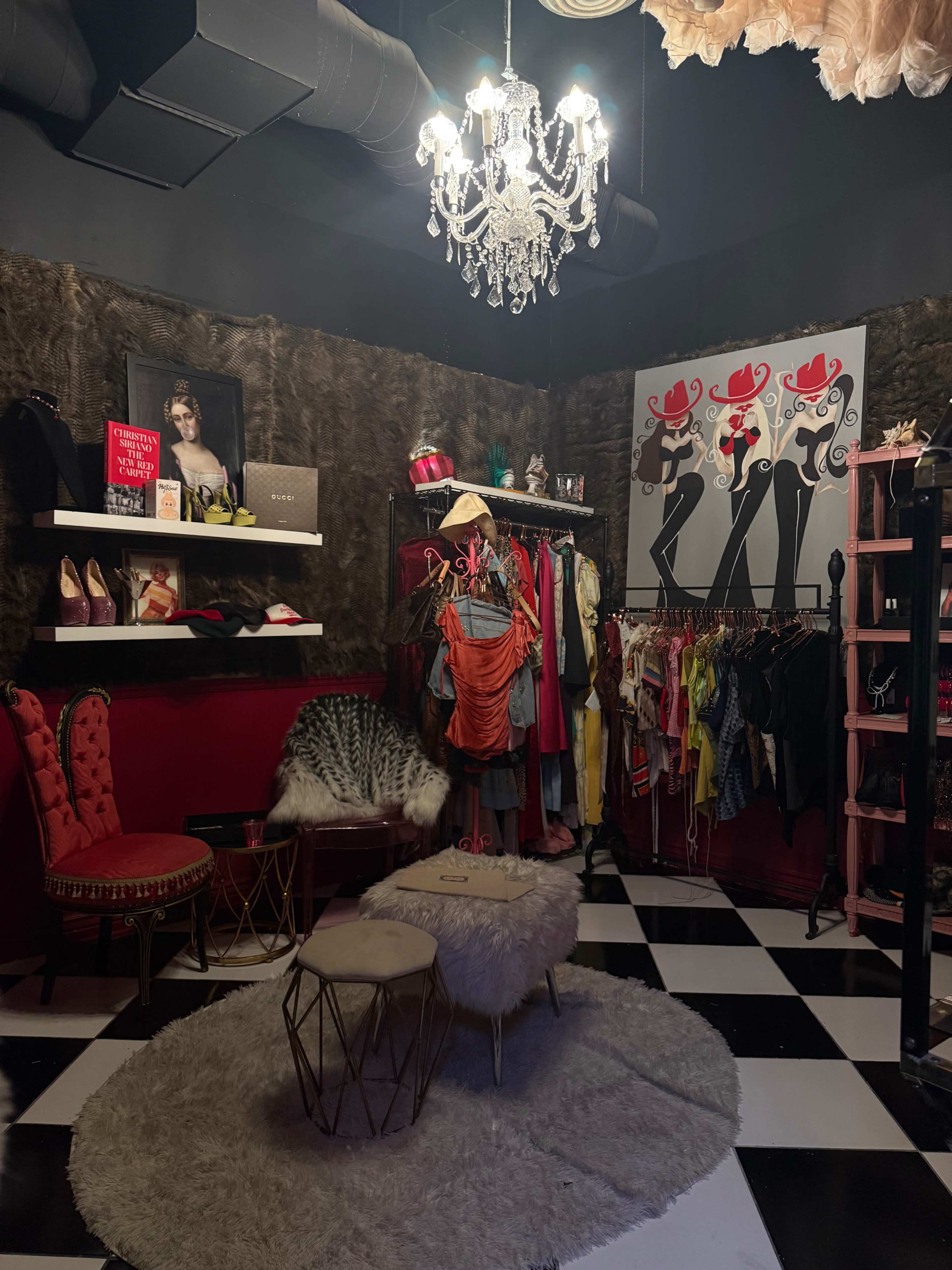 The image shows a boutique interior featuring a chandelier, a collection of clothing on racks, colorful high-heeled shoes on a shelf, and a plush seating area on a black and white checkered floor.
