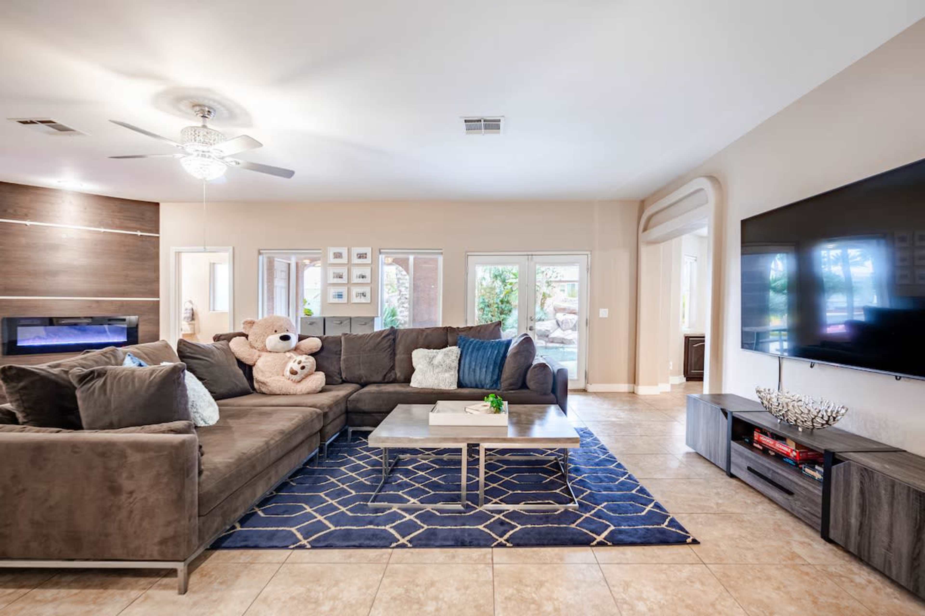 The image features a modern living room with a large sectional sofa, a coffee table, a television mounted on the wall, and a decorative rug on the floor.