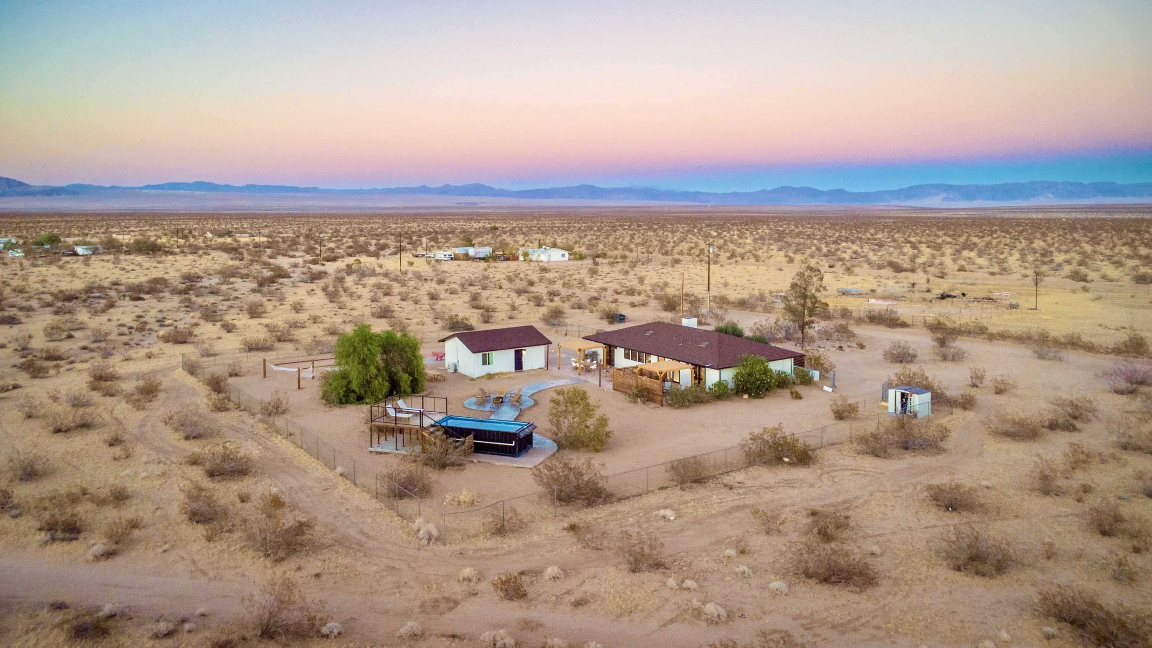The image shows a remote desert property featuring two buildings, a swimming pool, and sparse vegetation under a pastel-colored sky at dusk.