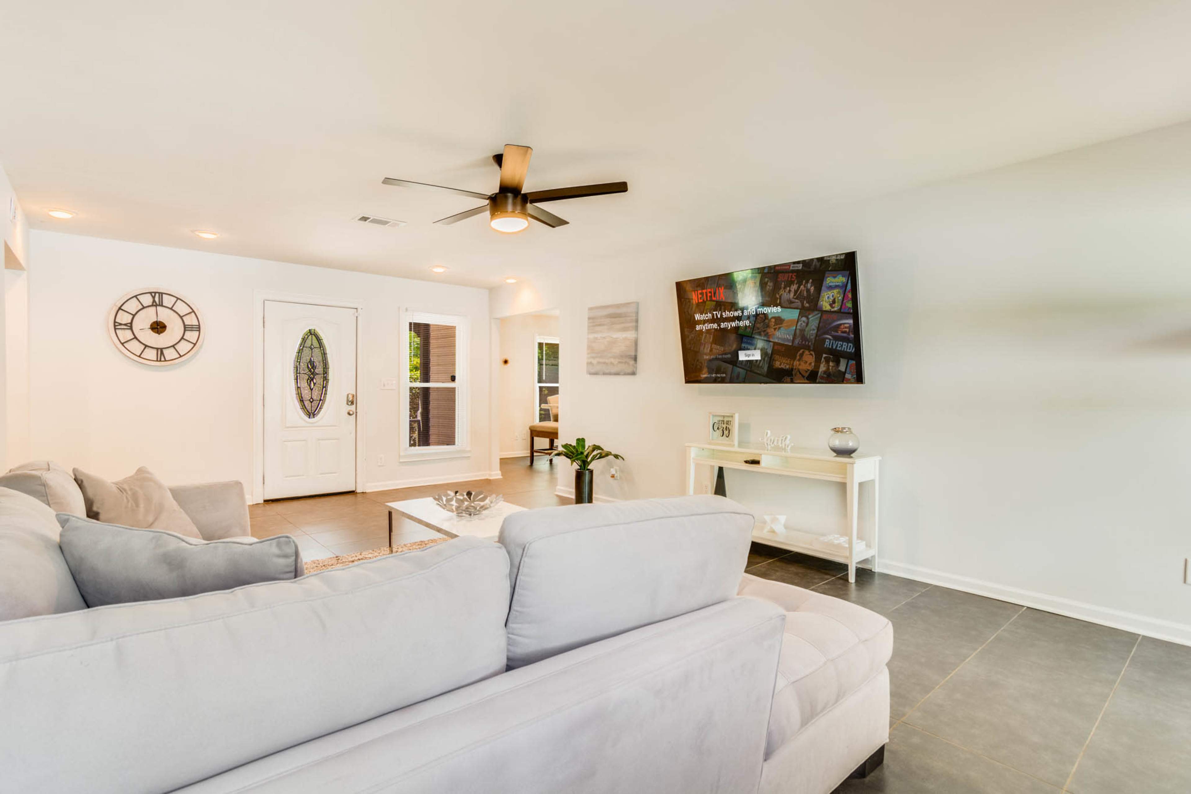 The image shows a bright living room featuring a light-colored sofa, a wall-mounted television, and a front door with a decorative window.