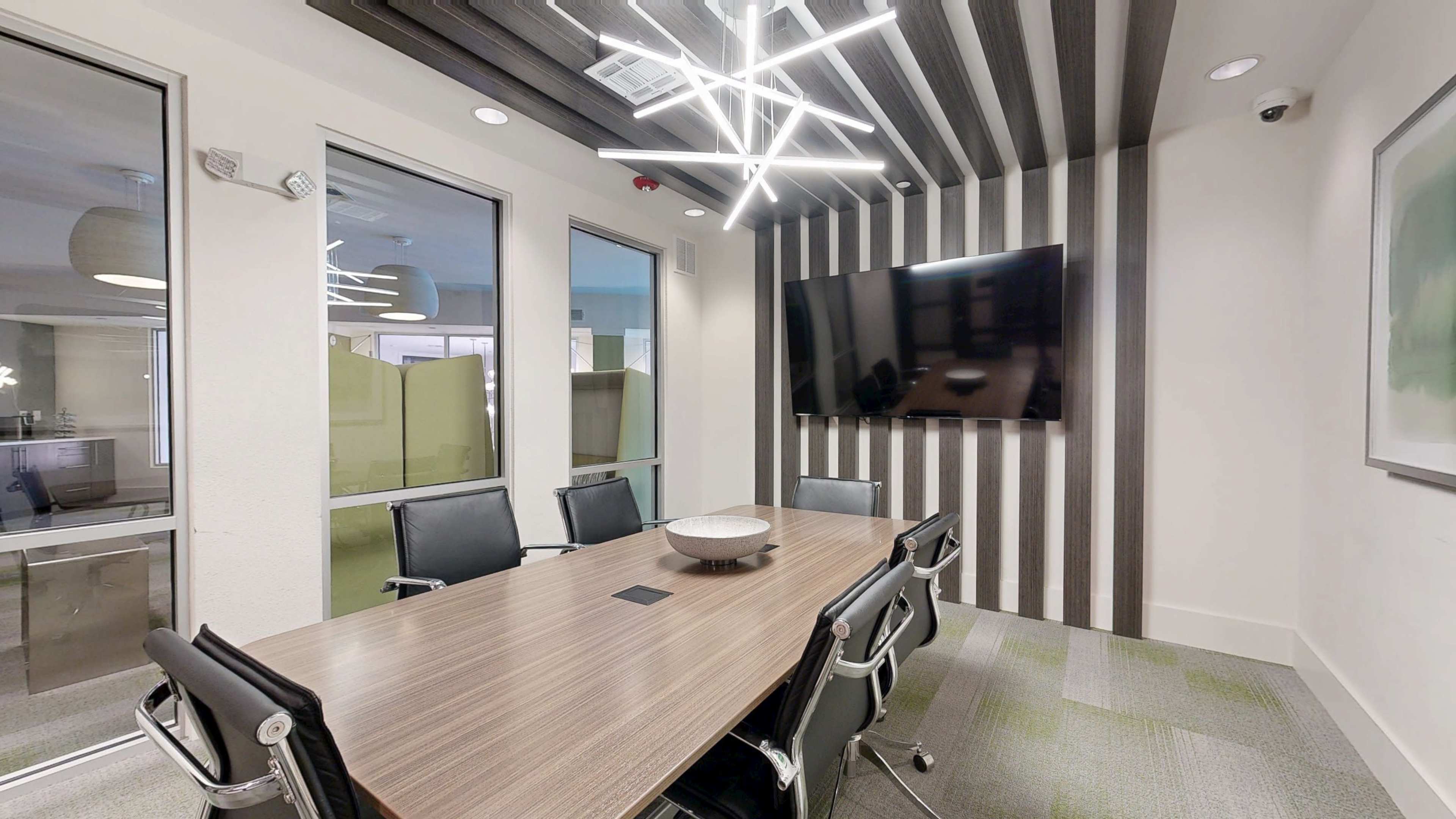 A modern conference room features a large wooden table surrounded by black chairs, with a flat-screen television mounted on a wall beneath contemporary lighting fixtures.