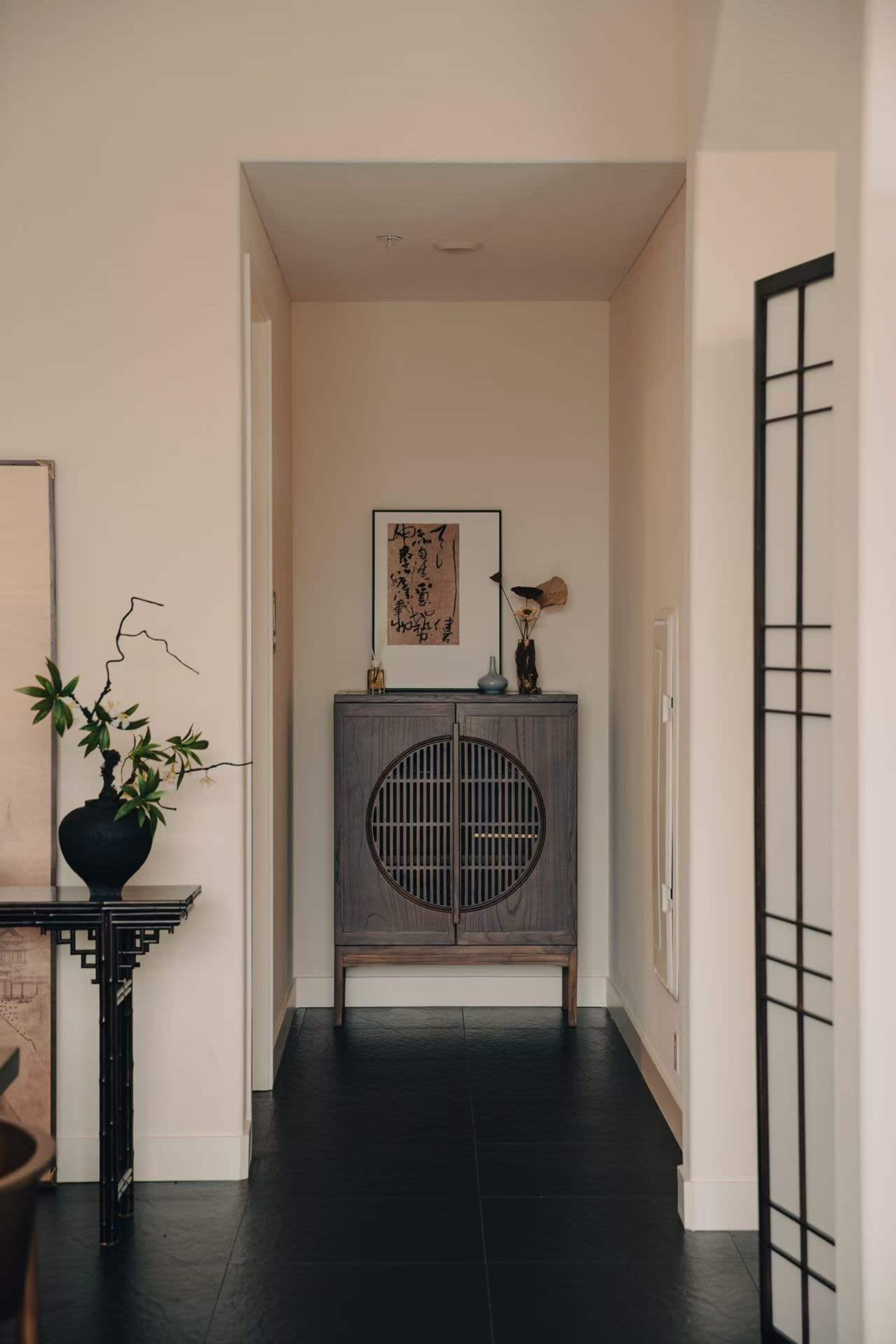 A narrow hallway features a dark wooden cabinet with circular doors, framed artwork on the wall, and a potted plant on a table.