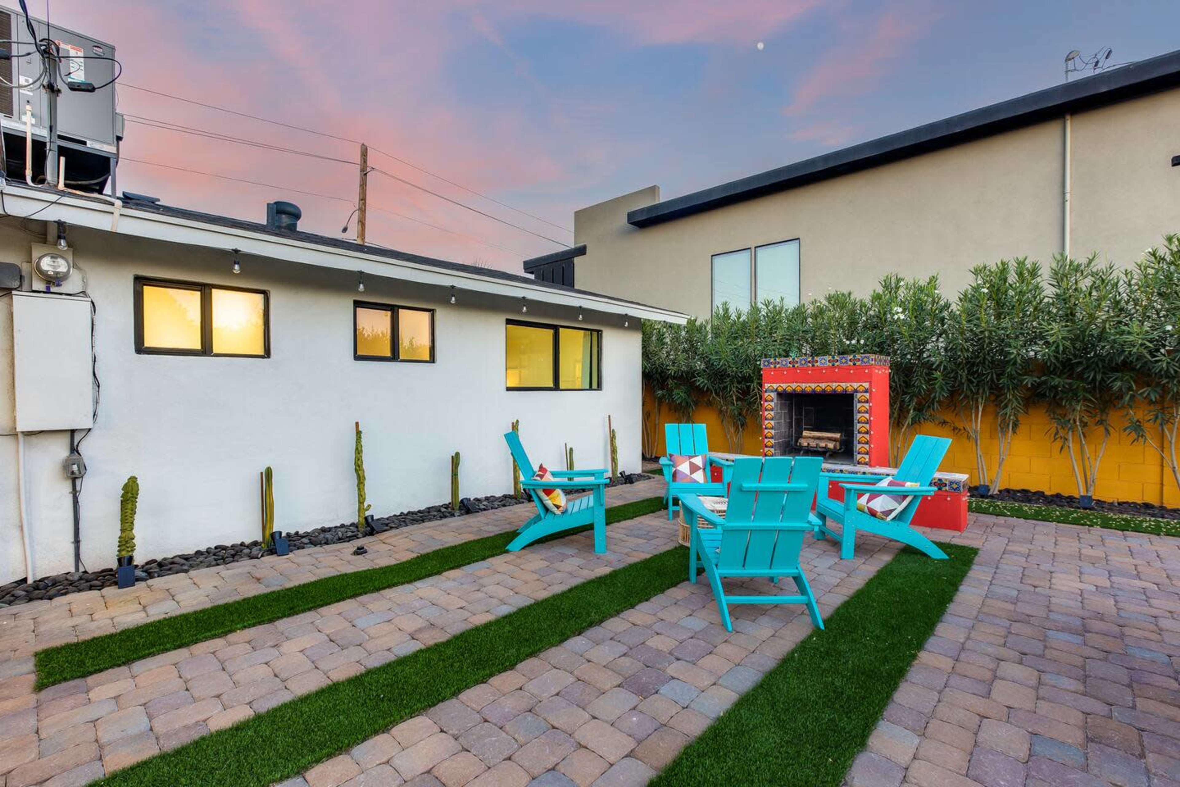 The image shows a small outdoor space with turquoise chairs arranged around a fire pit, surrounded by pavers and artificial grass, with a colorful outdoor fireplace against a wall.